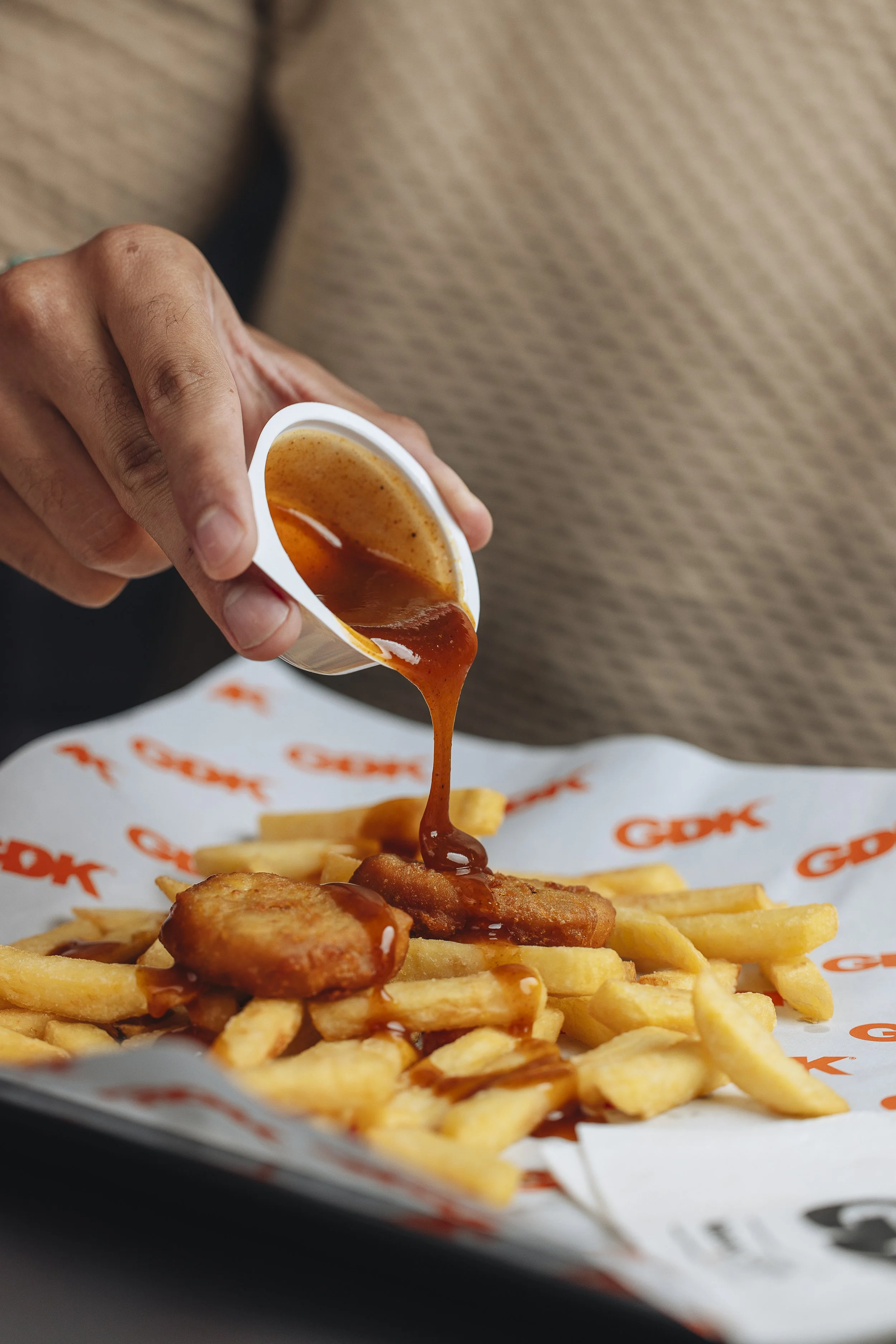 Hand pouring brown gravy over fried chicken and French fries at a tray lined with branded paper.