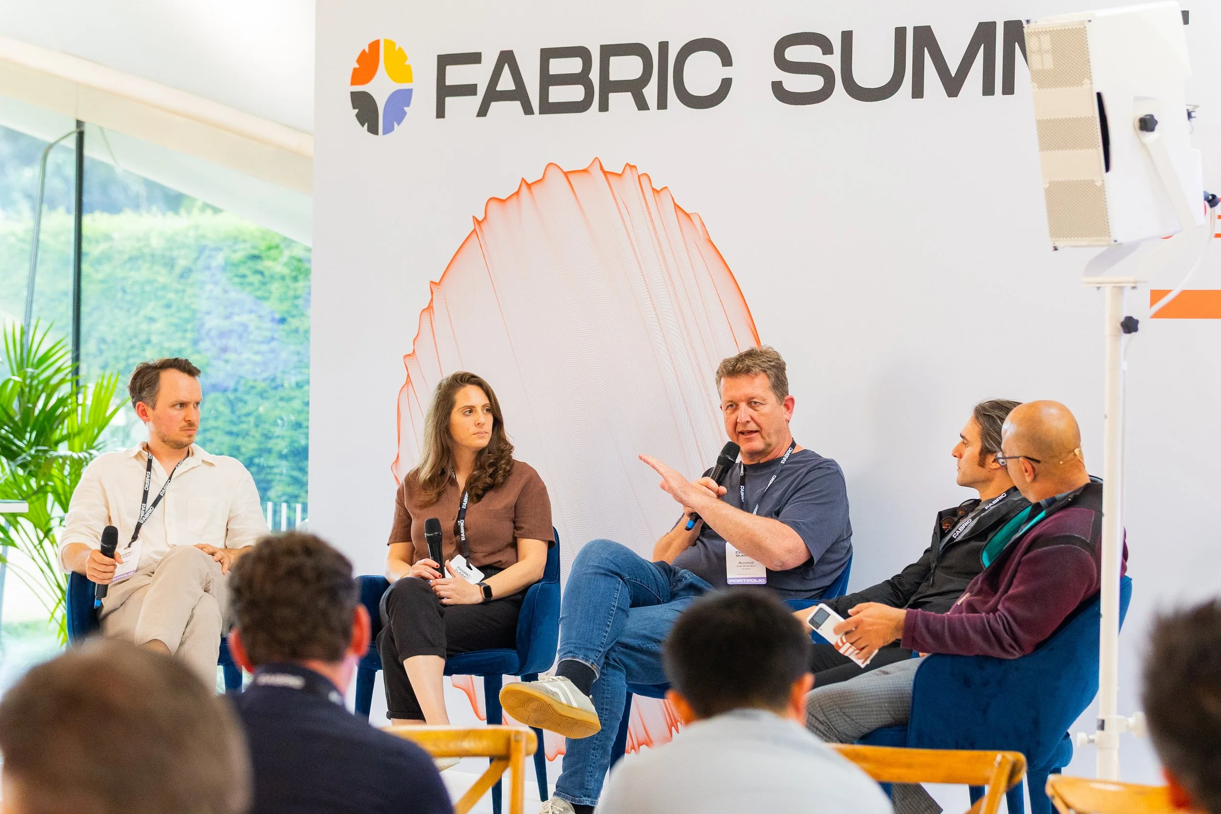 Panel discussion at Fabric Summit with five people on stage, four men and one woman, speaking and listening, with an audience in front, a large Fabric Summit banner behind them, and a plant to the side.