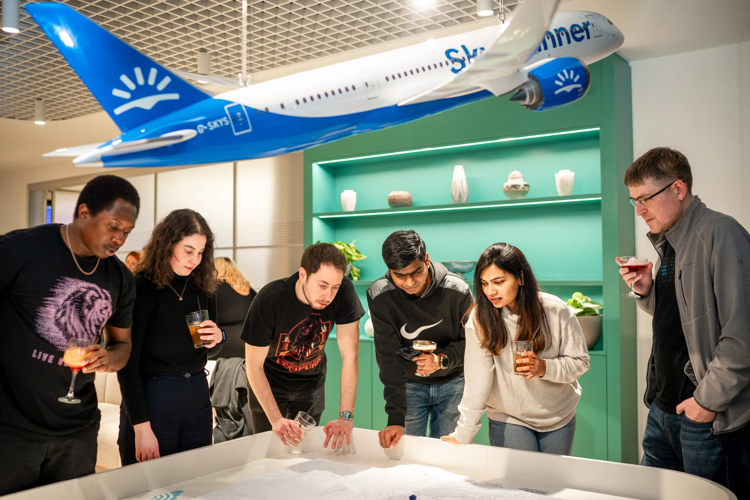 Group of six young adults gathered around a table looking at a large sand table or model display in an indoor space, with a blue and white airplane model hanging from the ceiling above them.