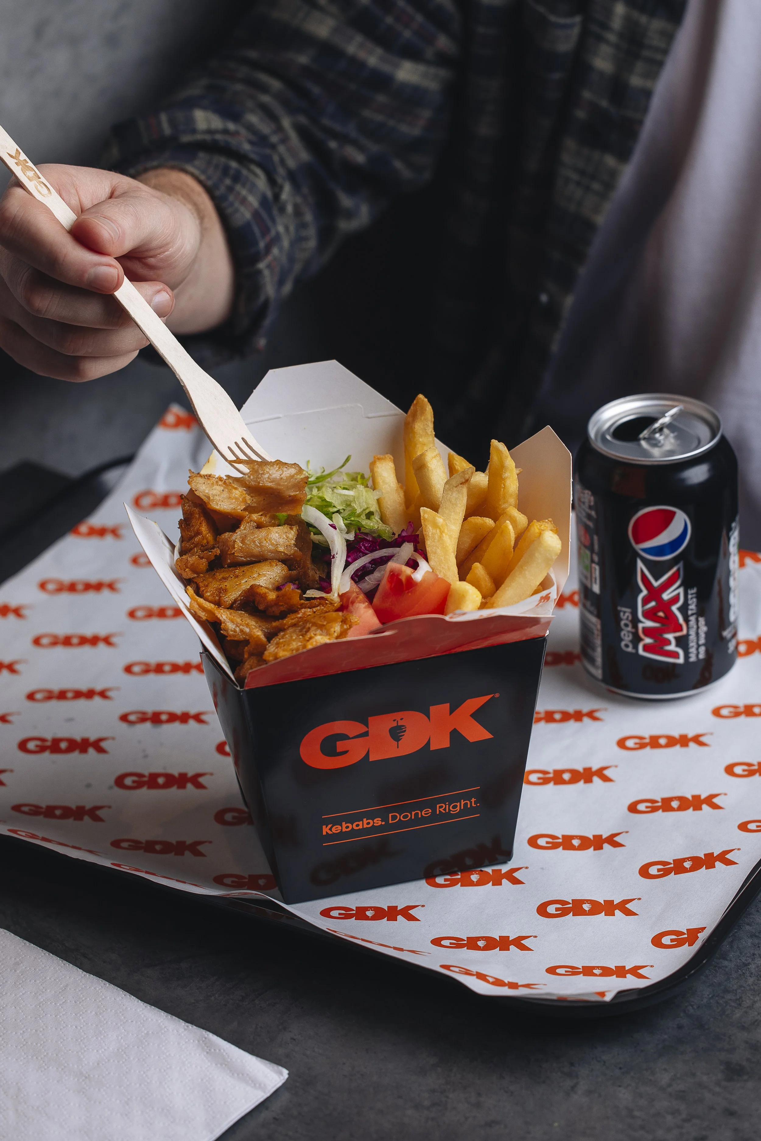 A person holding a wooden fork with fried chicken pieces in a GDK takeout box containing fries, lettuce, tomato, and onions, with a can of Pepsi Max on a GDK-branded tray.