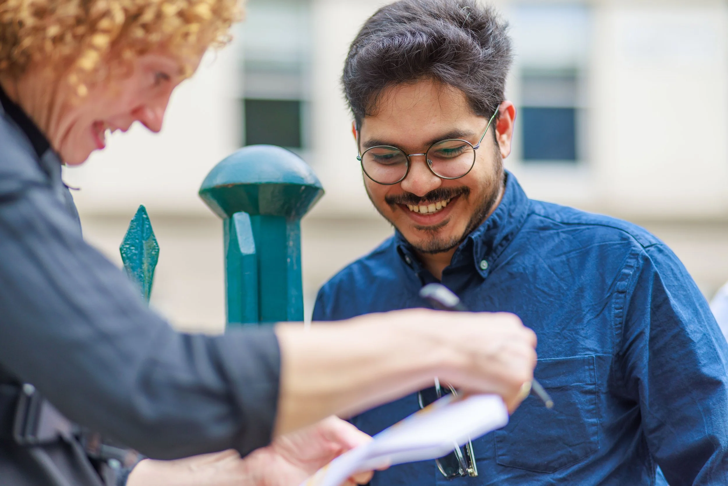 Two people, a woman and a man, are smiling and looking at a notebook or clipboard outdoors near a green metal railing. The woman has curly red hair and the man has glasses and dark hair.