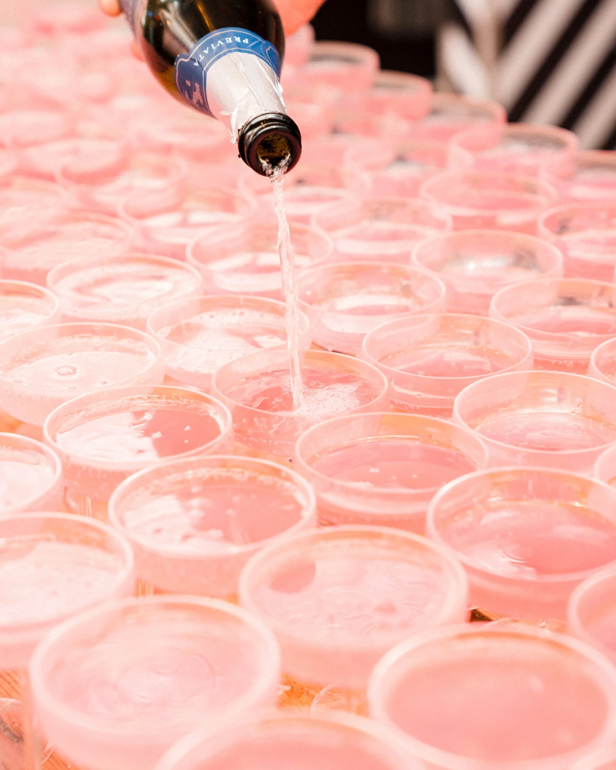 A bottle pouring clear liquid into small pink plastic cups arranged on a table.