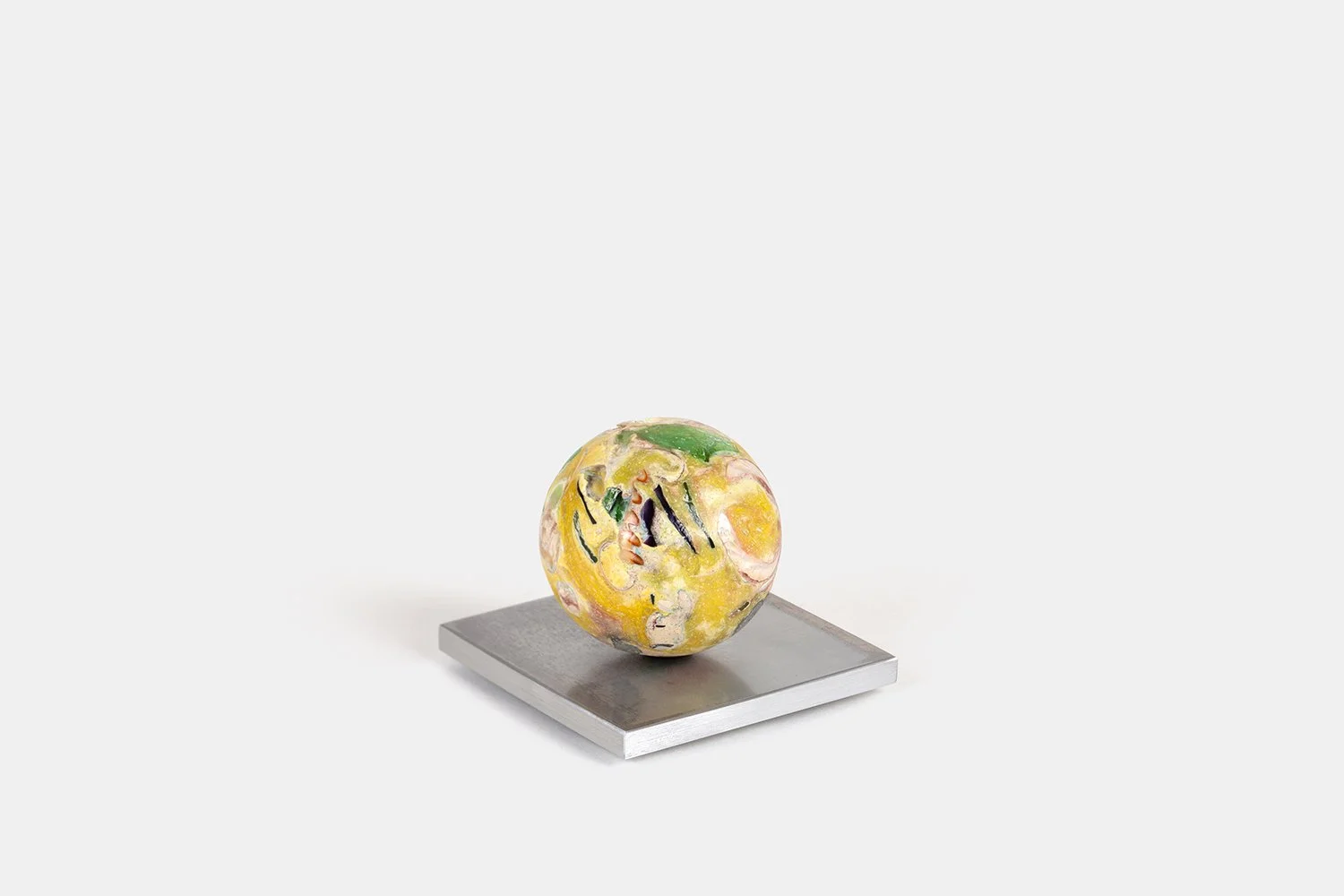 Yasue Maetake Orb Materia—Yuzu, 2026. Compact yellow spherical form with green sea glass and dark linear marks, resting on an angled square aluminum base.