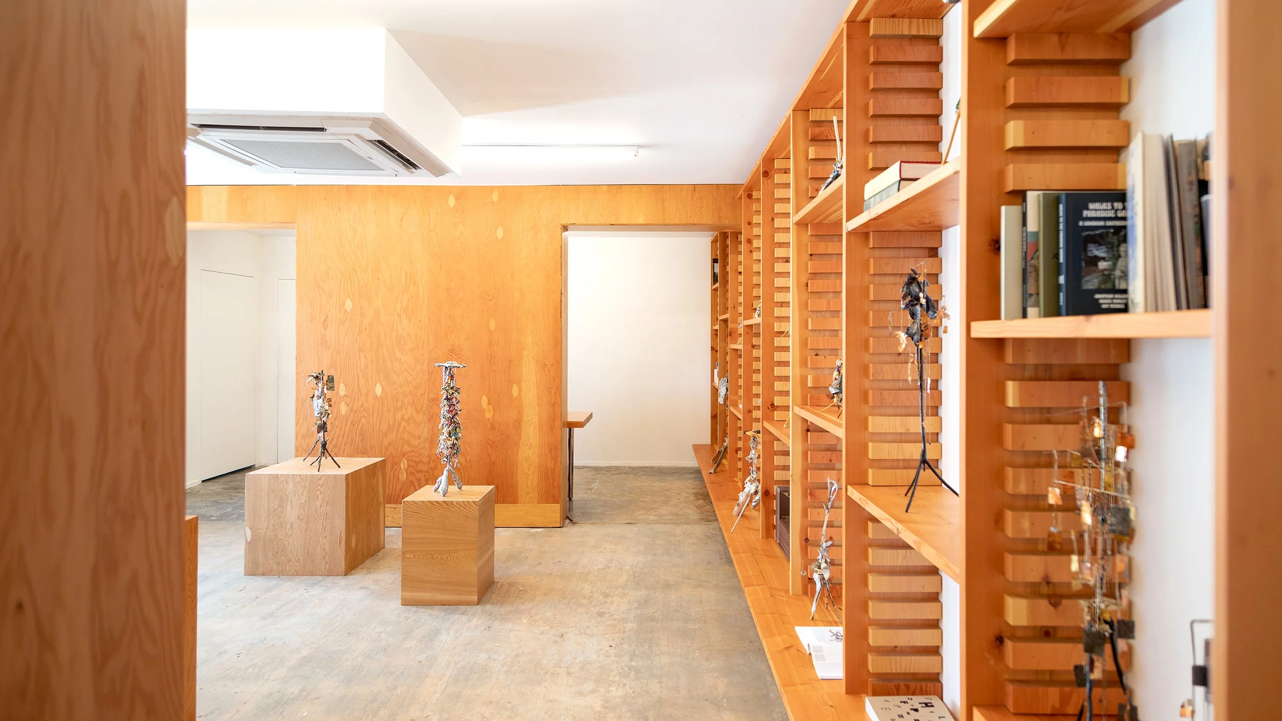 Yasue Maetake Three-Legged Idols gallery installation: view through wooden doorway into gallery with two sculptures on wooden pedestals and tall bookshelf of small three-legged works on the right