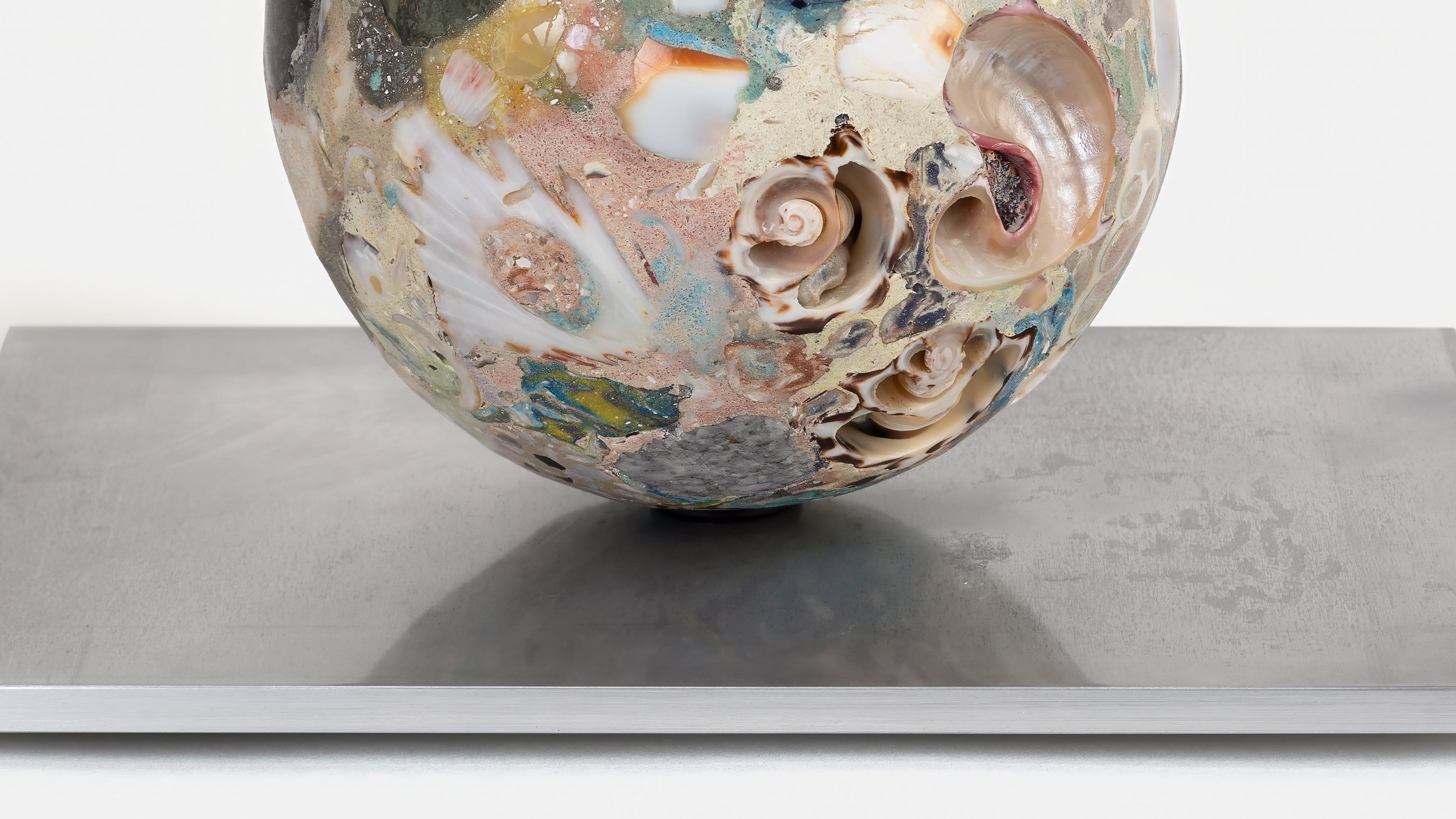 Yasue Maetake Orb Materia—Carved Algos detail: close-up of a carved sphere densely embedded with spiral shells and organic fragments, resting on a rectangular aluminum base