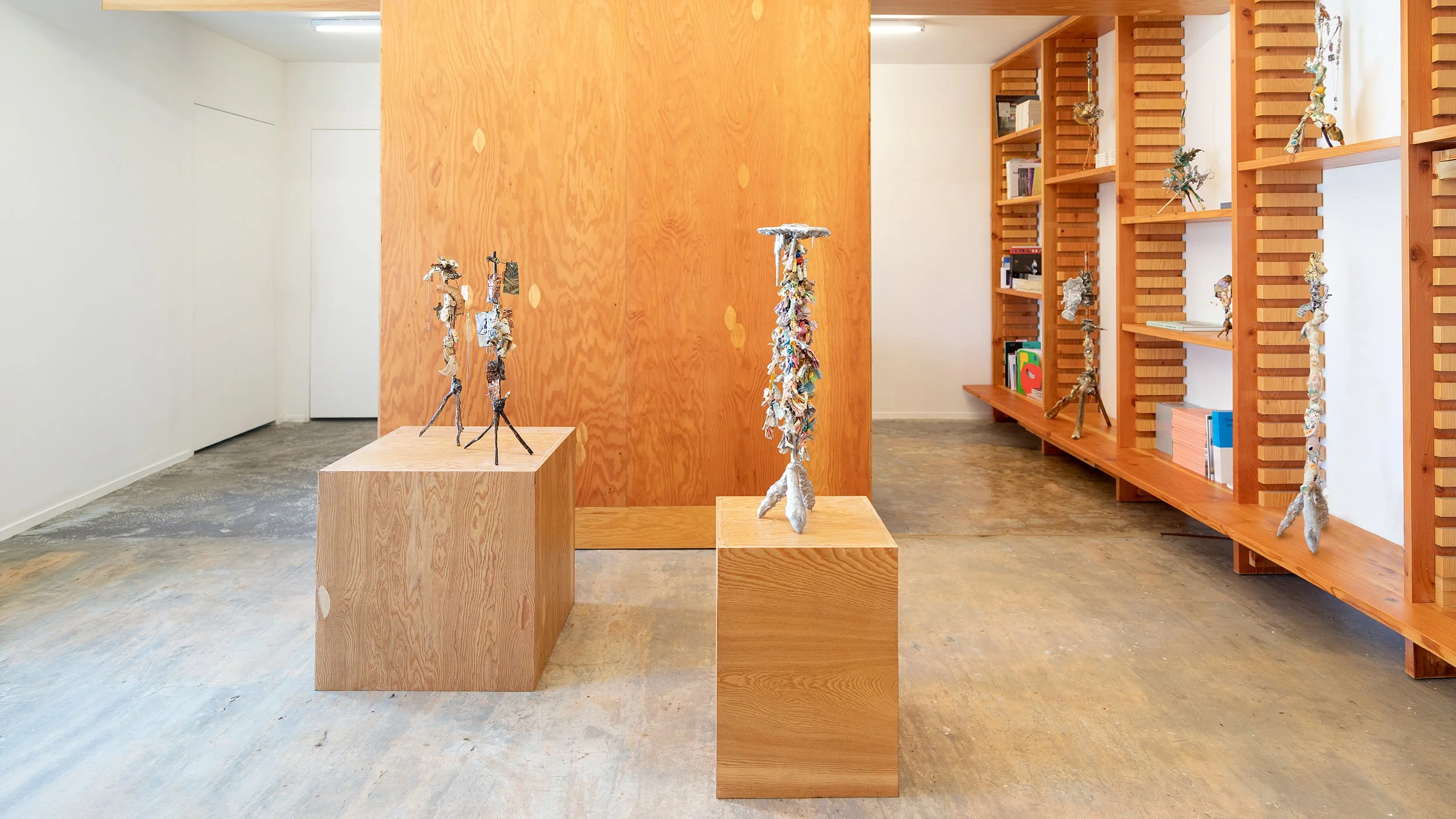 Yasue Maetake Three-Legged Idols gallery installation: two wooden pedestals with three-legged sculptures in the foreground, tall wooden bookshelf of additional works on the right in warm gallery space