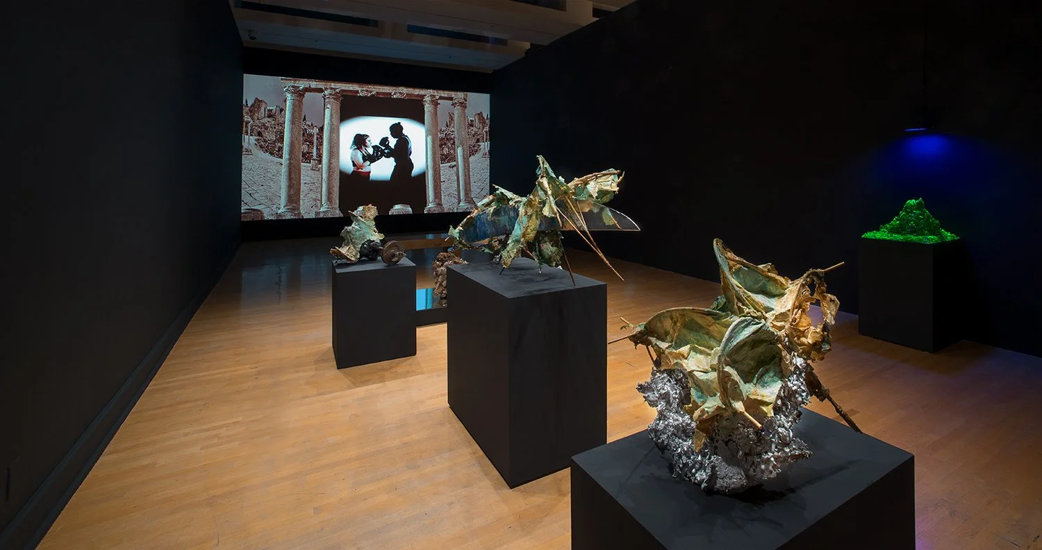 Art gallery featuring metallic insect sculptures on black pedestals, with a projection screen displaying a silhouette of a couple and ancient ruins in the background.