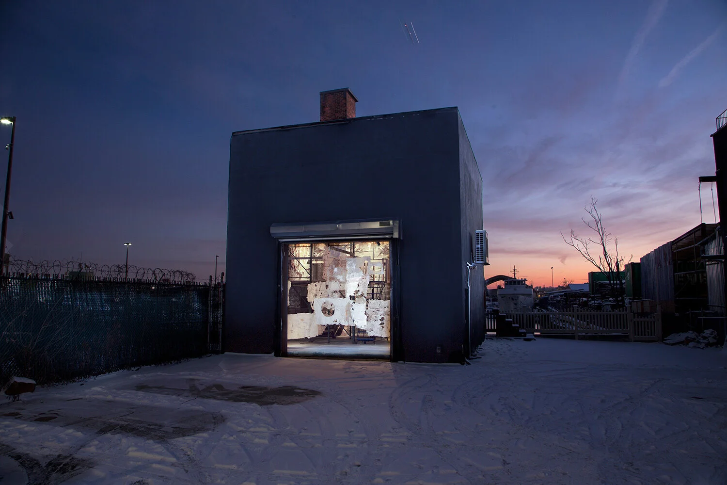 A small black building with an open garage door showing an lit interior, against a sunset sky. Snow covers the ground outside and a fence with barbed wire is on the left.