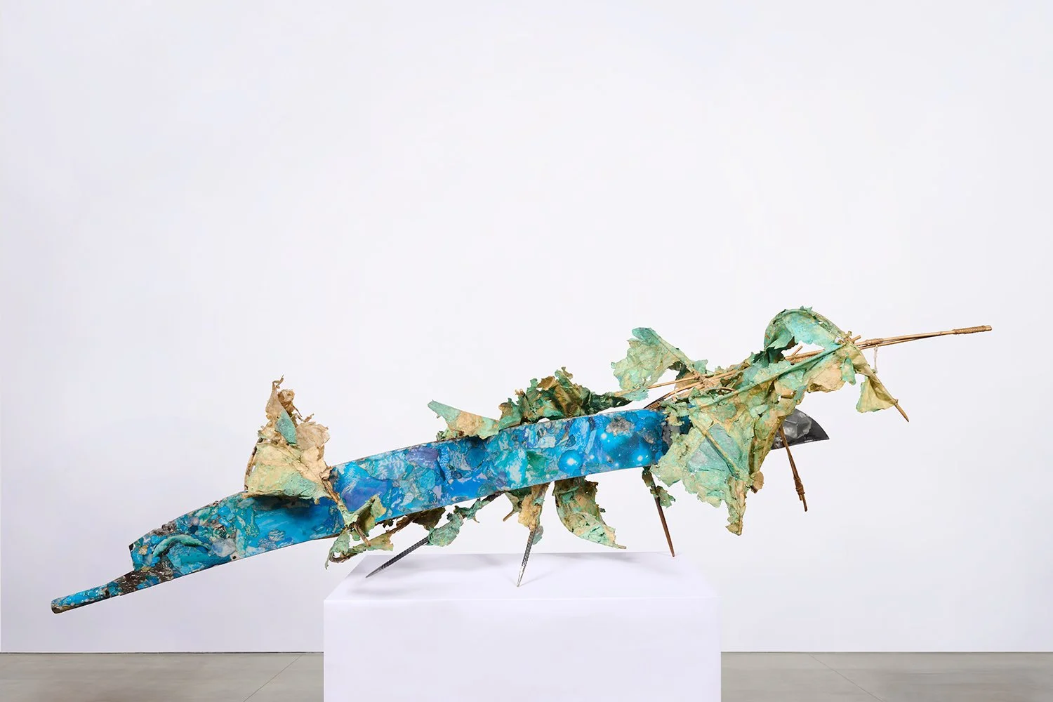 Mixed-media sculpture resembling a dragonfly, composed of painted metal, paper, and organic materials, displayed on a white pedestal against a plain white wall.