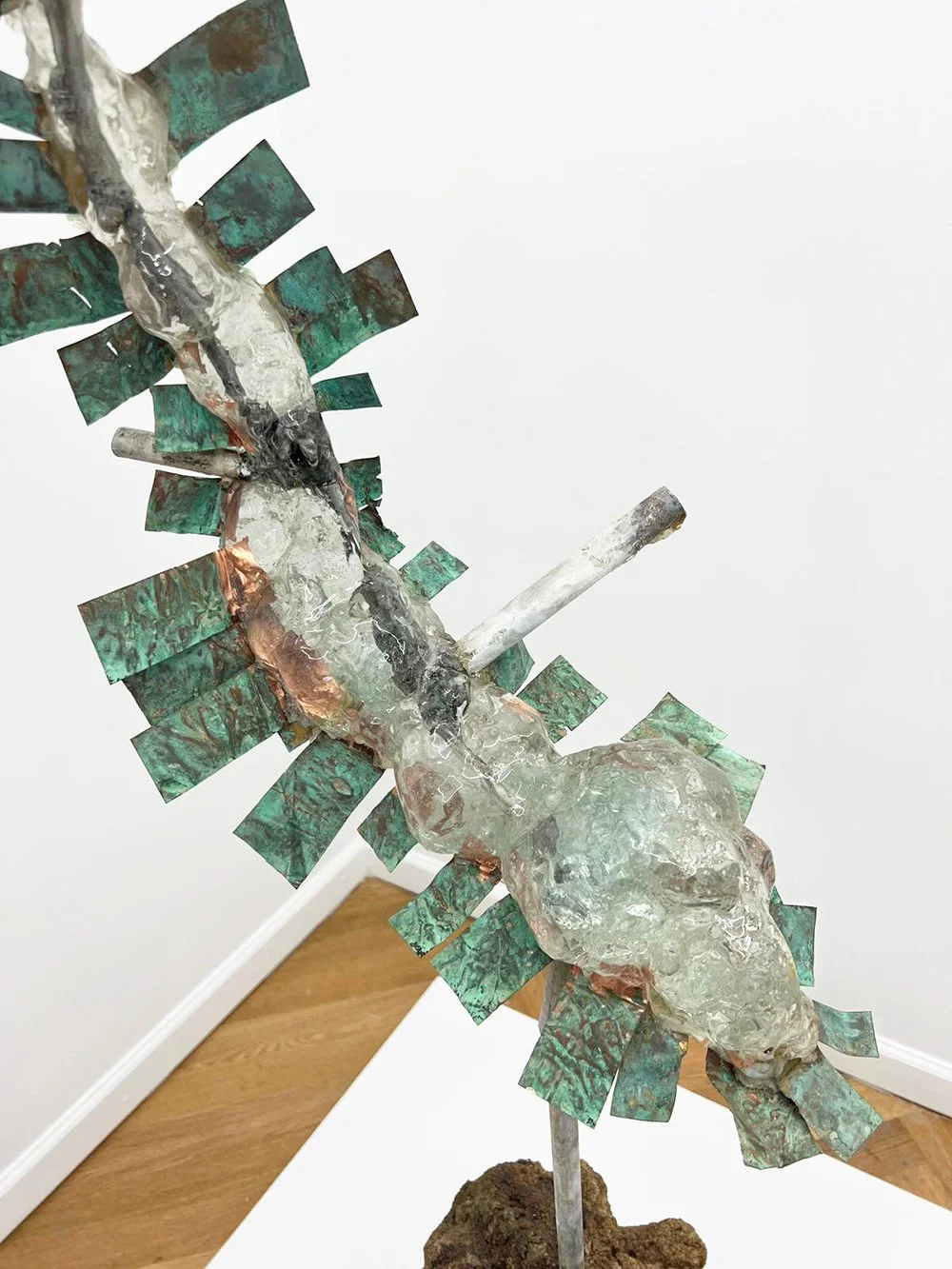Abstract sculpture resembling a bird with metallic and textured surfaces, green and copper patina accents, mounted on a base of soil or clay.