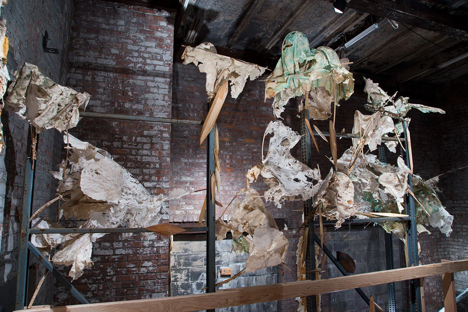 An art installation of large, weathered paper and fabric sculptures mounted on metal frames against a red brick wall, with a wooden railing in the foreground.