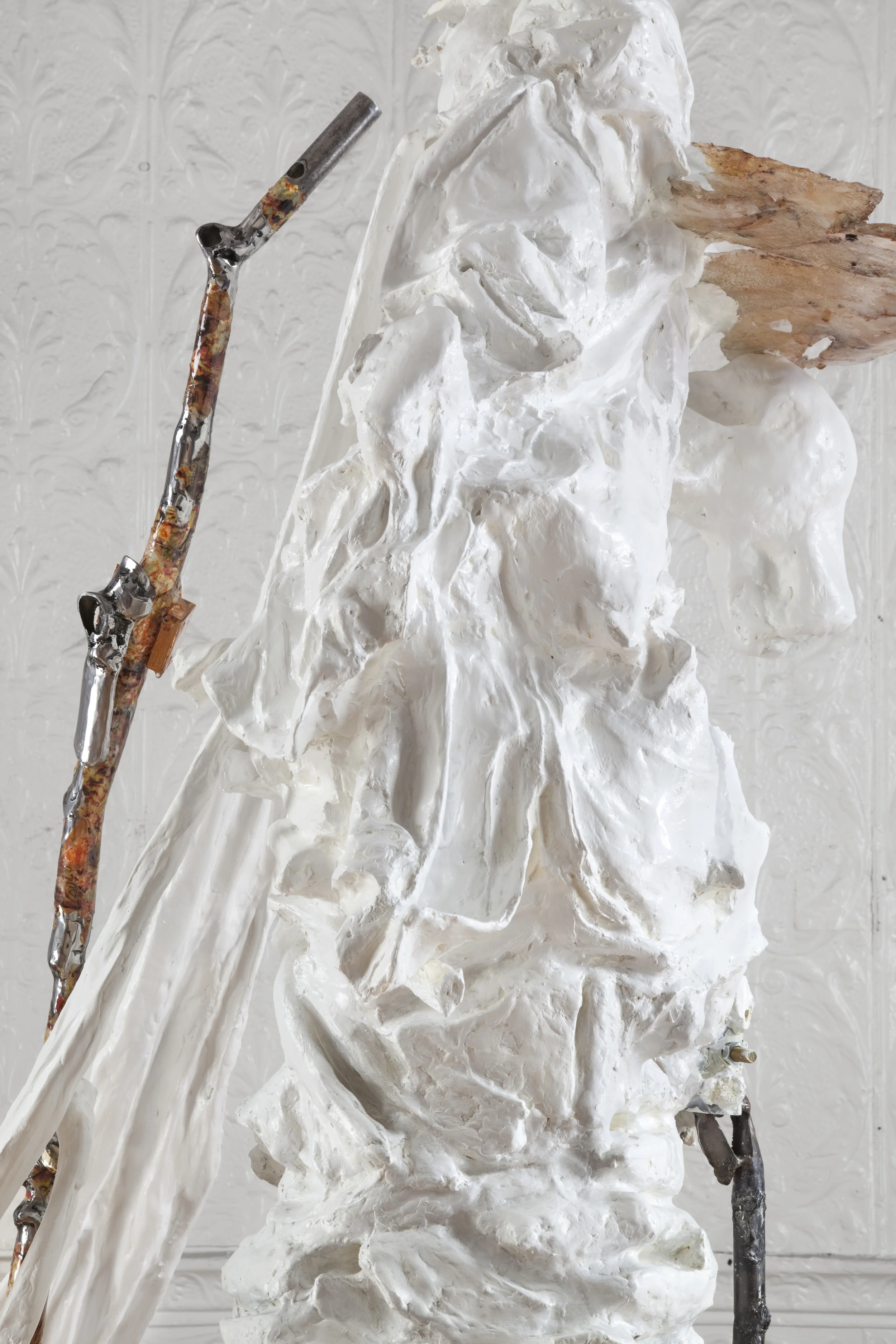 An artist's sculpture of a face. The sculpture is partially covered in white plaster or clay, with some areas still unformed. A wooden and metal armature supports the sculpture in front of a plain, textured white wall.