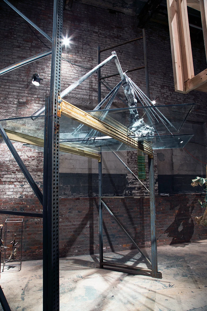 An artistic installation featuring glass panels, metal frames, and wires mounted on a brick wall with industrial lighting, creating shadows and reflections.