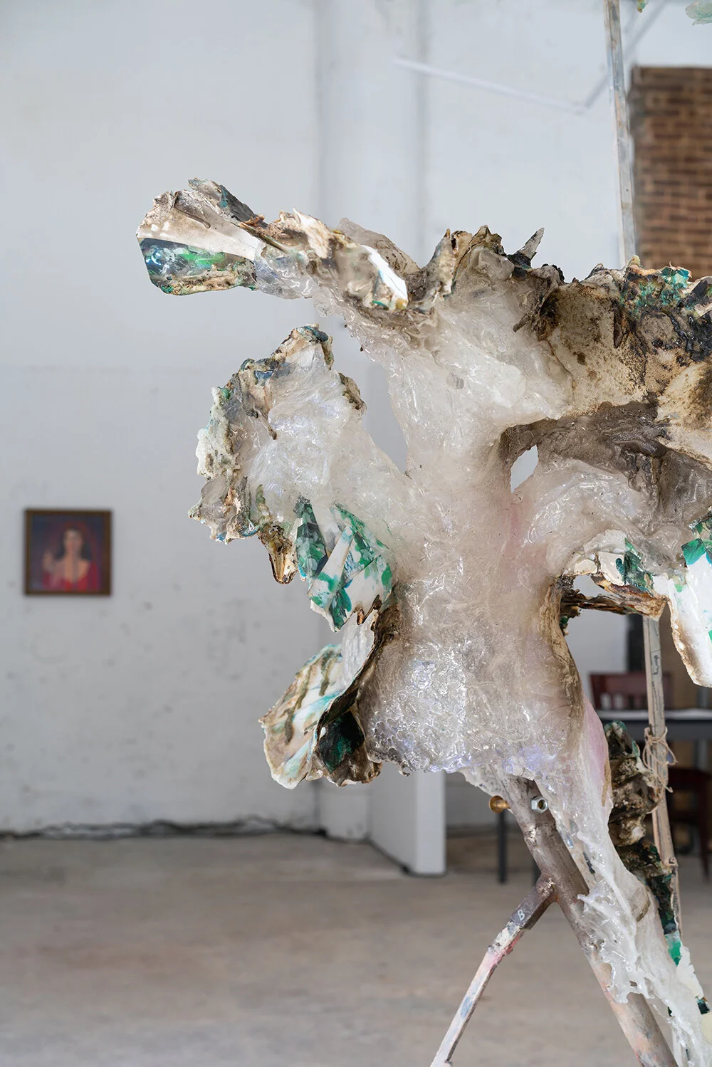 Abstract sculpture made of mixed materials including glass and metal in an art gallery, with a brick wall and artwork in the background.