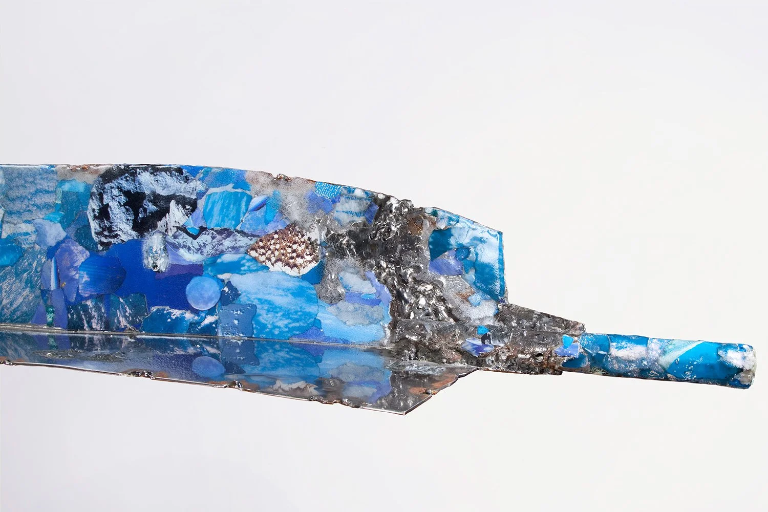 Close-up of Bullet Turquoise (2023) with a metallic sculpture resembling a knife with a blue mosaic pattern, against a plain white background.