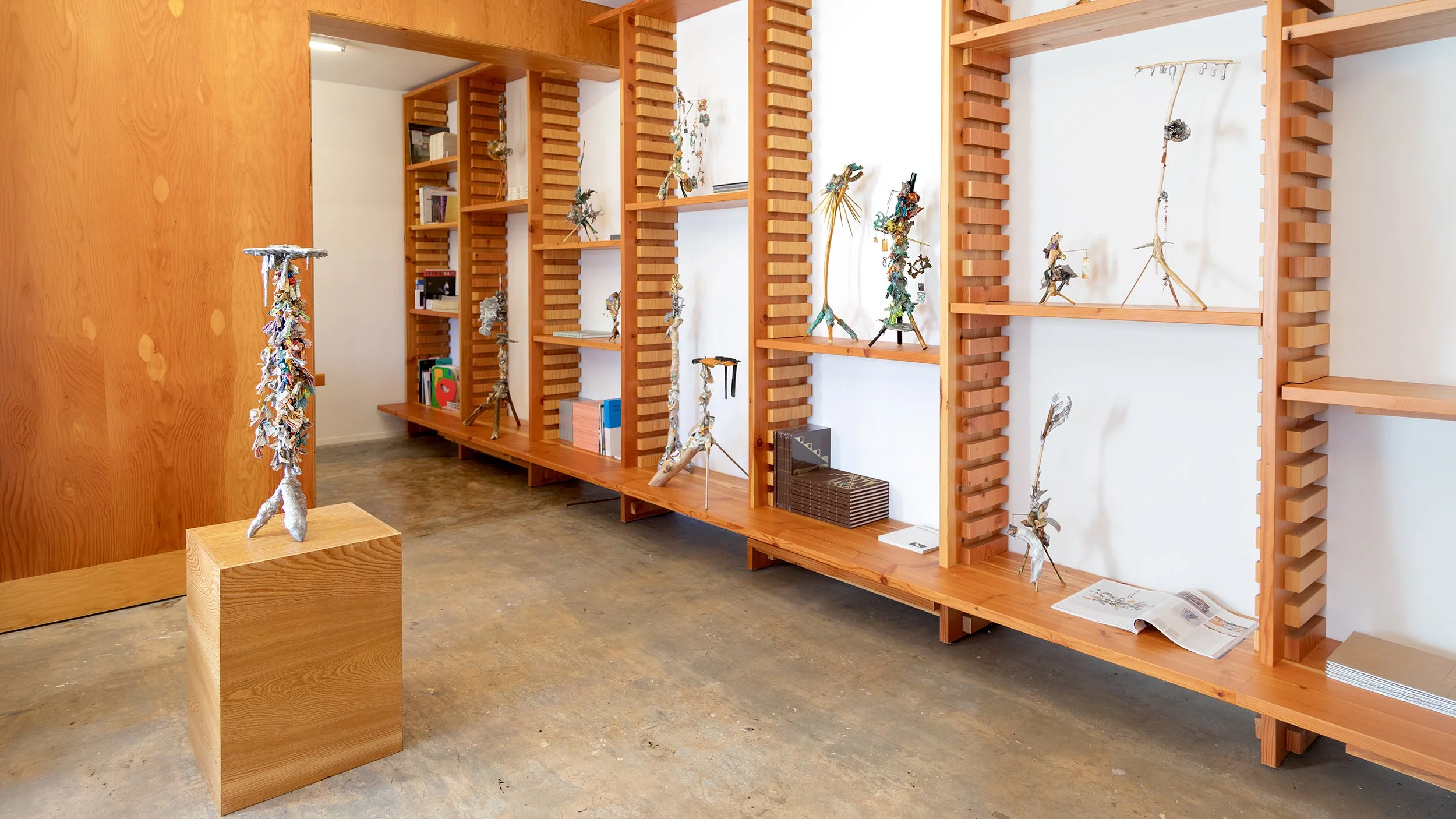 Yasue Maetake Three-Legged Idols gallery installation: wooden bookshelf section with several small three-legged sculptures placed among books on multiple shelves
