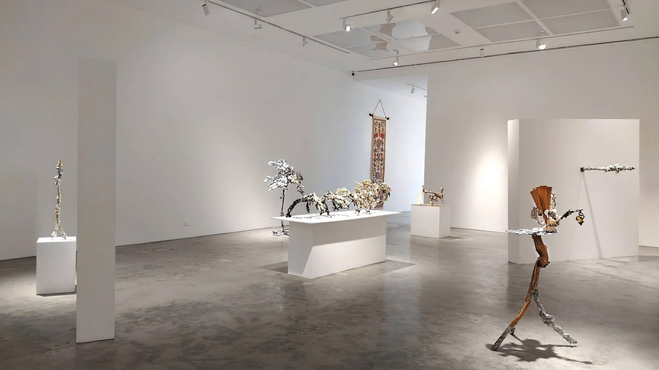 Yasue Maetake Mare Fecunditatis wide gallery view: tall silvery tree-like on floor left, long pedestal with multicolored dangling forms center, small beige on pedestal right front, human-scale orange-brown tripod piece right back in white space