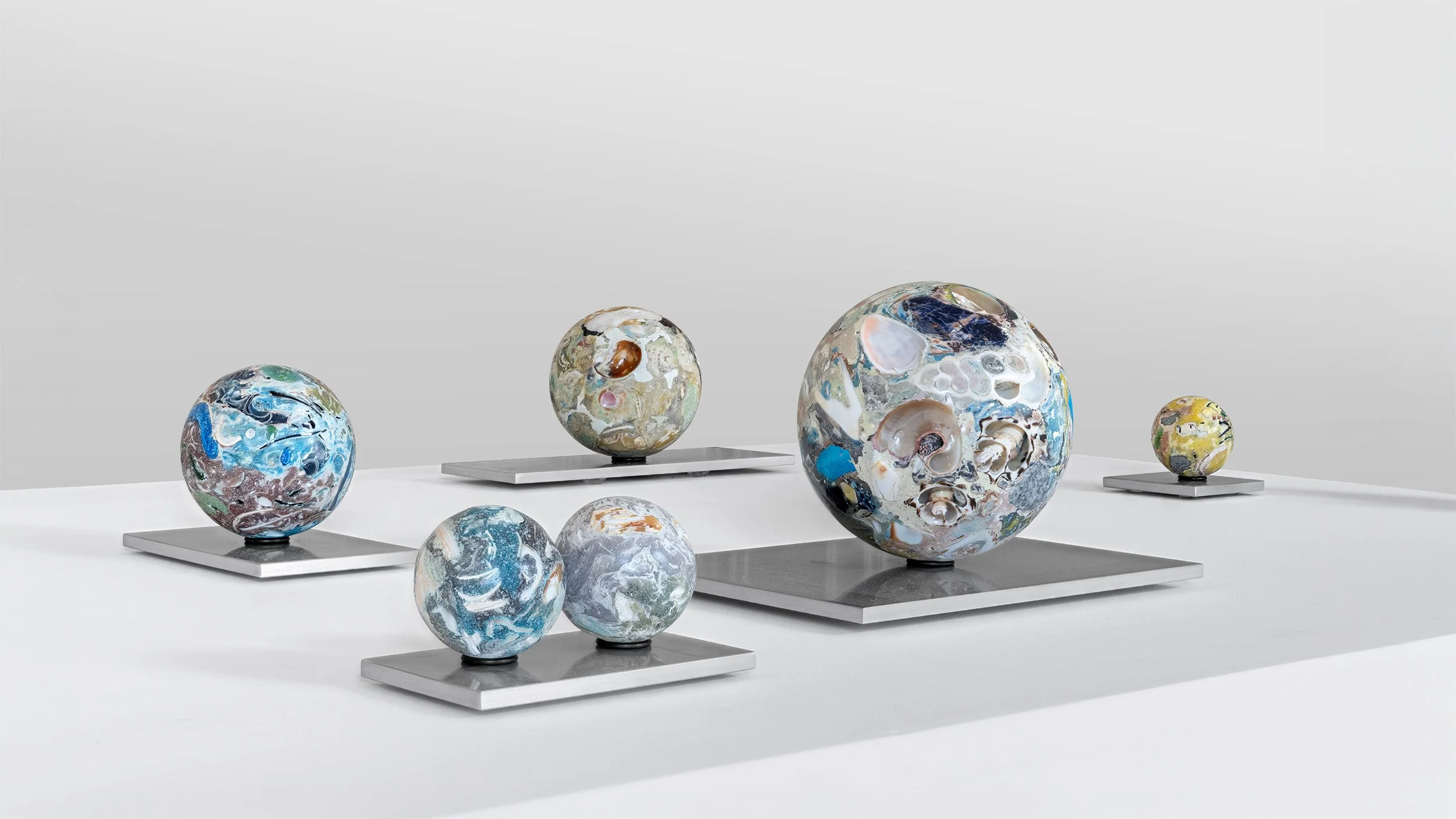 Yasue Maetake Orb Materia—Carved Algos: group of multicolored carved spheres of varying sizes on aluminum bases, with a large detailed sphere as the focal point against a plain light background