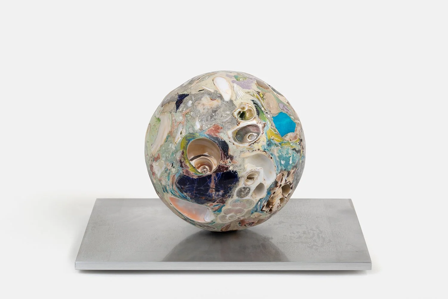 Yasue Maetake Orb Materia—Mayfly, 2026. Dense spherical form with deep shell cavities, spiral openings, and mixed blue, beige, and dark mineral surfaces on a rectangular aluminum base.