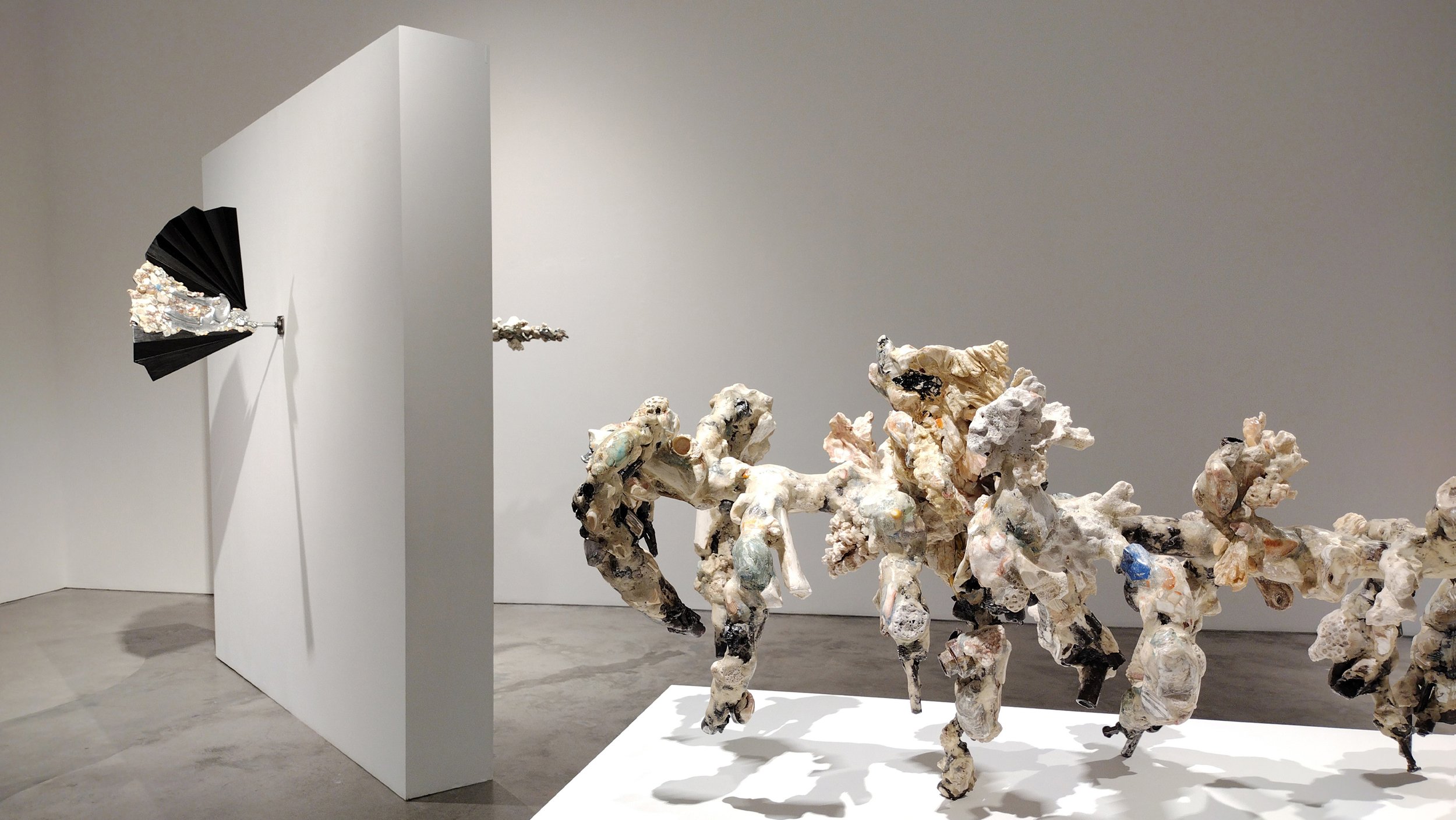 Yasue Maetake Mare Fecunditatis gallery view: black fan-shaped piece wall-mounted on panel left, long pedestal with row of multicolored dangling forms center in white space