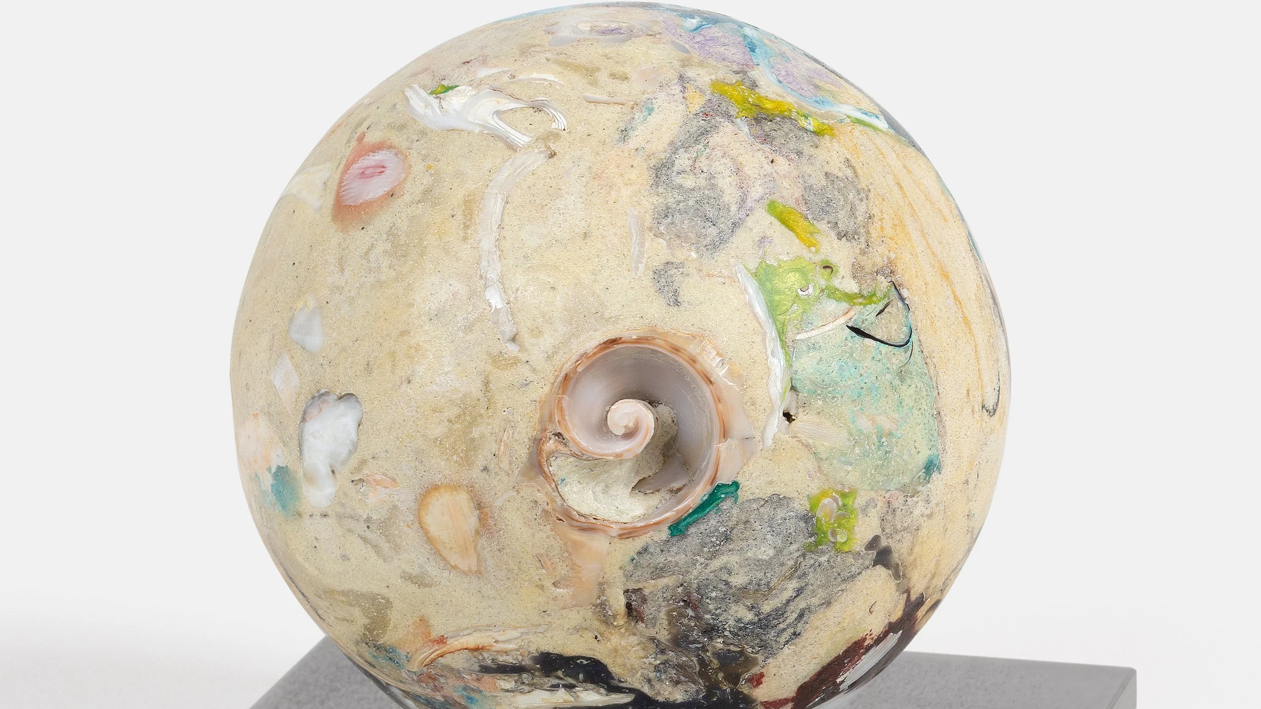 Yasue Maetake Orb Materia—Carved Algos detail: close-up of a carved sphere with a prominent spiral shell embedded in a sandy surface of multicolored mineral and organic fragments