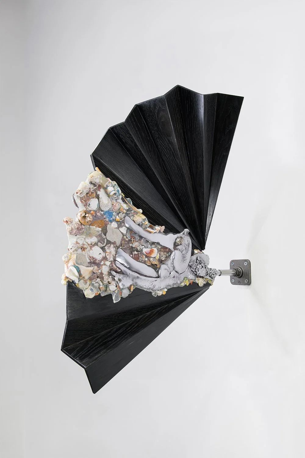 Yasue Maetake, Ceremonial Delta I, 2025. Wall-mounted hybrid sculpture featuring a large black pleated wooden fan structure with a dense, protruding organic form of multicolored shells, stones, and metal.