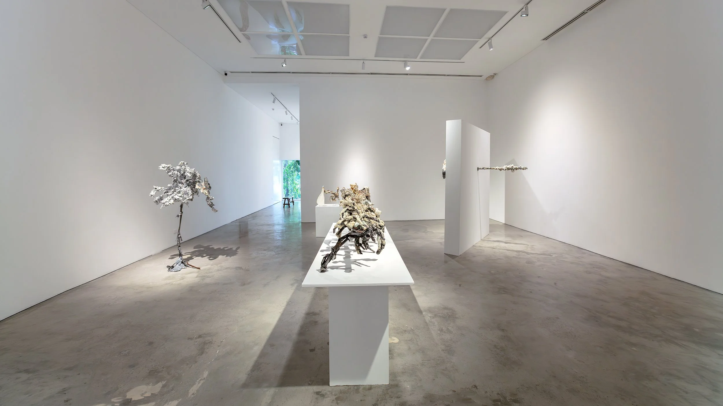 Yasue Maetake Mare Fecunditatis exhibition view: tall silvery crystalline tree-like structure on floor left, human-scale clustered form on pedestal center, long horizontal bar wall-mounted right in white gallery with green door background
