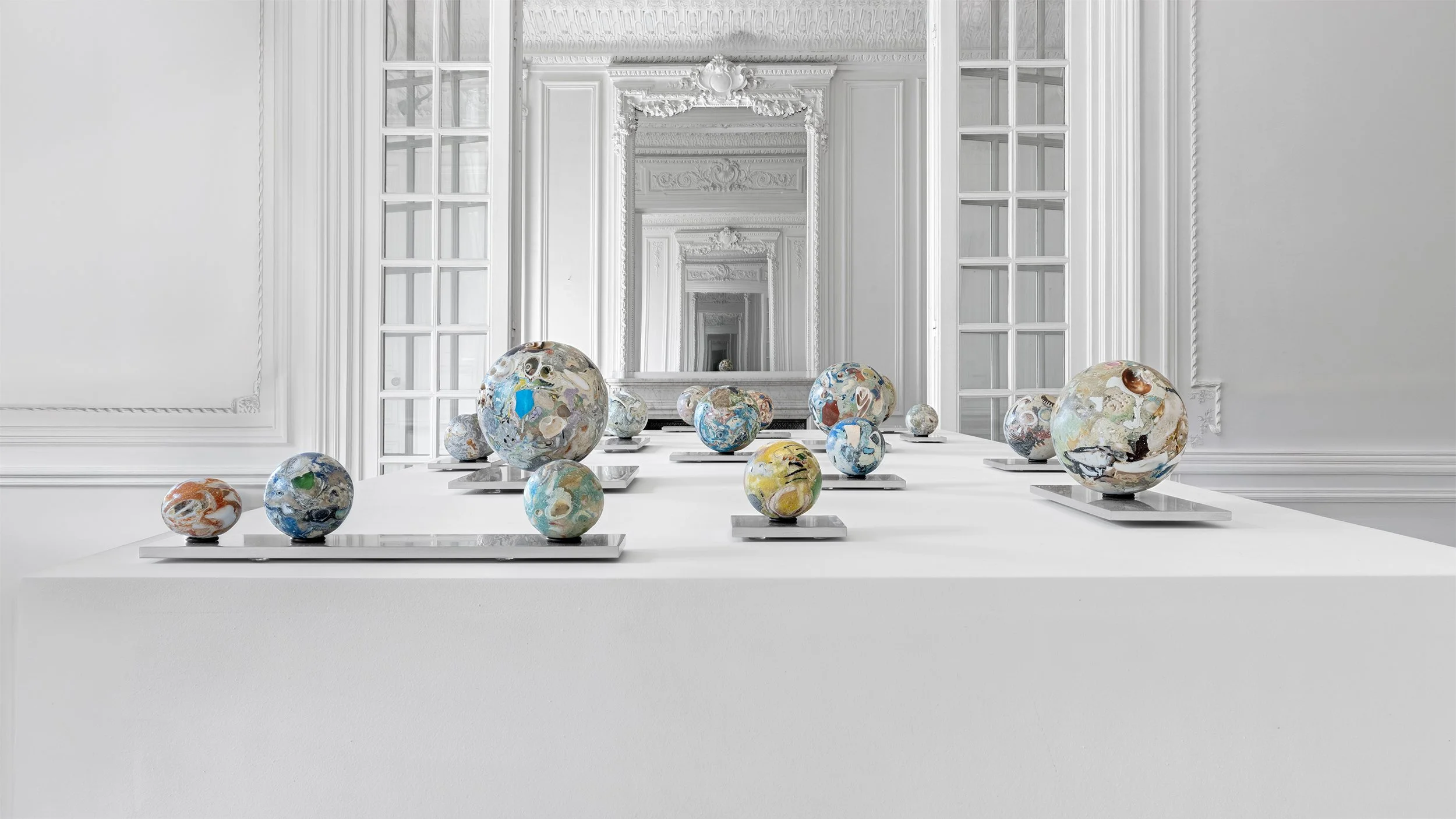 Yasue Maetake Orb Materia—Carved Algos gallery installation: long view of multicolored carved spheres arranged on a white platform with infinite mirror reflections in a European classical white room