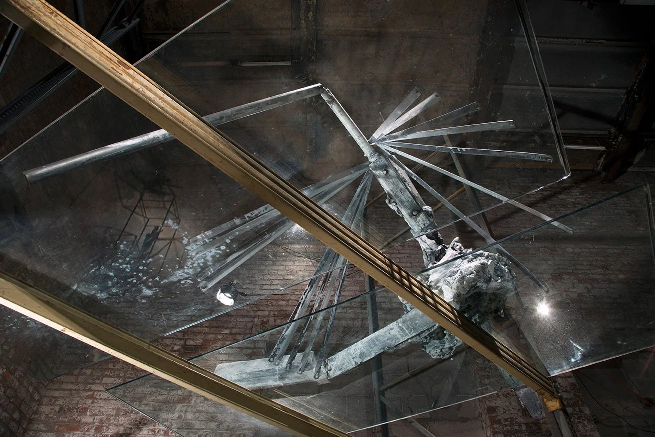 Broken glass display case with shattered glass fragments and metal structures inside, mounted on a brick wall in an industrial setting.