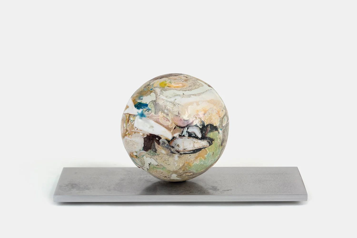 Yasue Maetake Orb Materia—Cocoon, 2026. Spherical form with layered beige, white, and pale green mineral surfaces containing multiple shell fragments, resting on a long rectangular aluminum base.