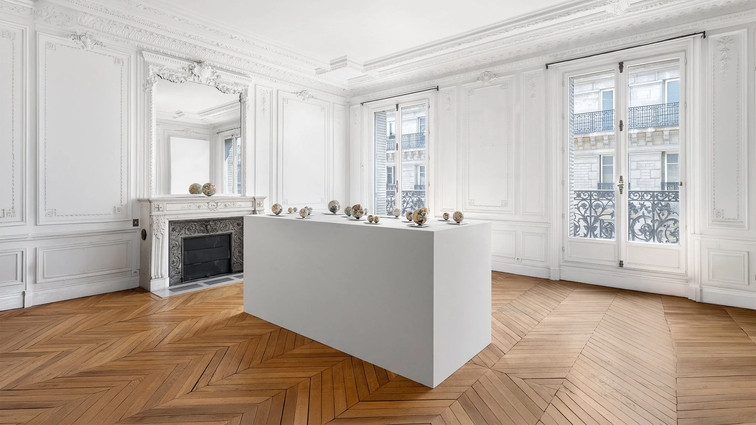 Yasue Maetake Orb Materia—Carved Algos gallery installation: wide angled view of multicolored carved spheres on a central white platform in a European classical room with fireplace, tall windows, and wooden floor