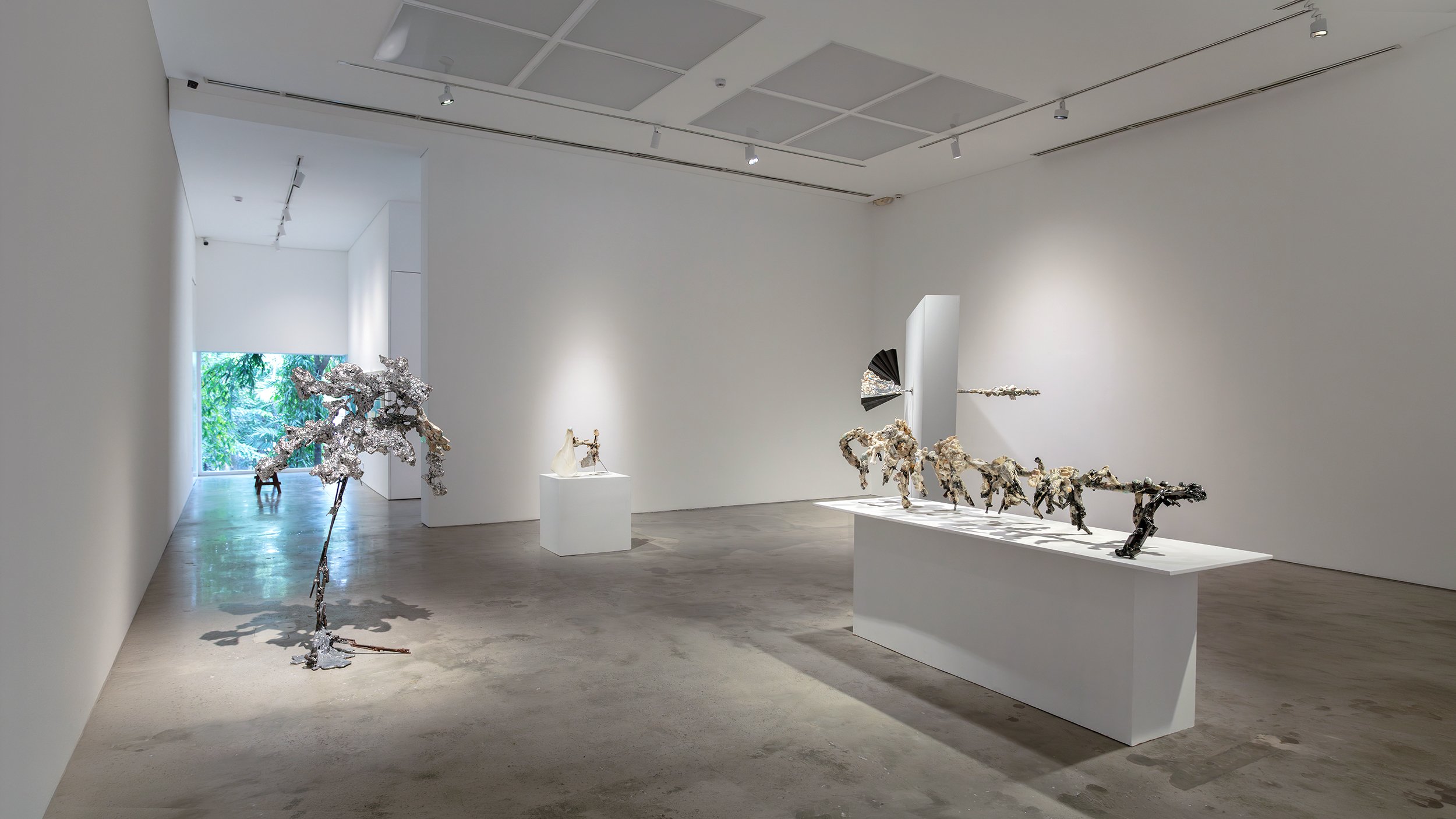 Yasue Maetake Mare Fecunditatis gallery installation: tall silvery crystalline branch on floor left, small white form on cube pedestal center, row of black twisted sculptures on shelf right in white space