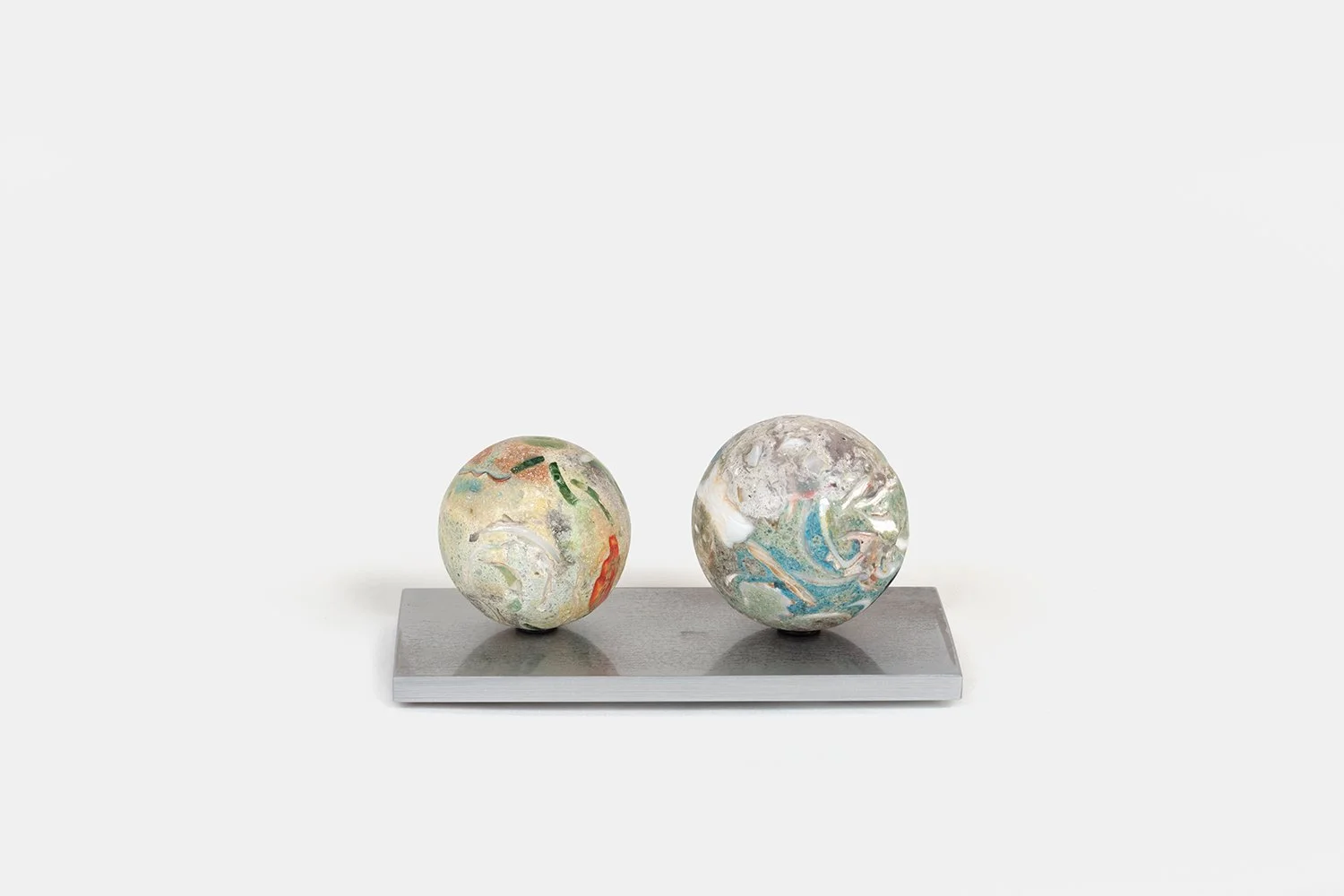 Yasue Maetake Orb Materia—Binoculars, 2026. Two spherical sculptures on a rectangular aluminum base, the left with warm orange and green tones and the right with cooler blue, white, and green mineral surfaces.