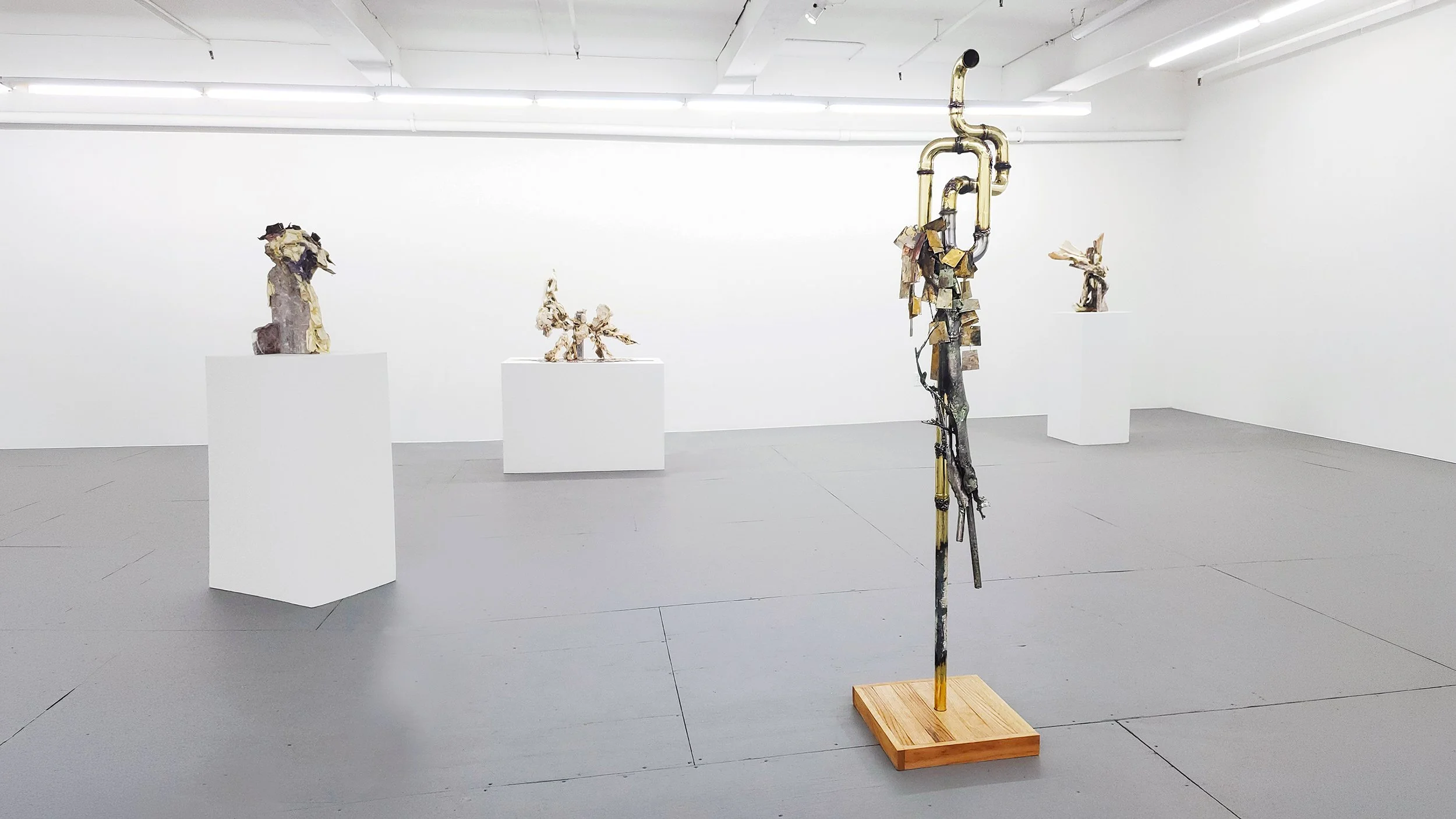 Yasue Maetake Transmutation gallery installation: tall non-representational sculpture of brass pipes, metal fragments, and organic elements on a wooden base in the foreground, with other bone and metal sculptures on white pedestals in the background