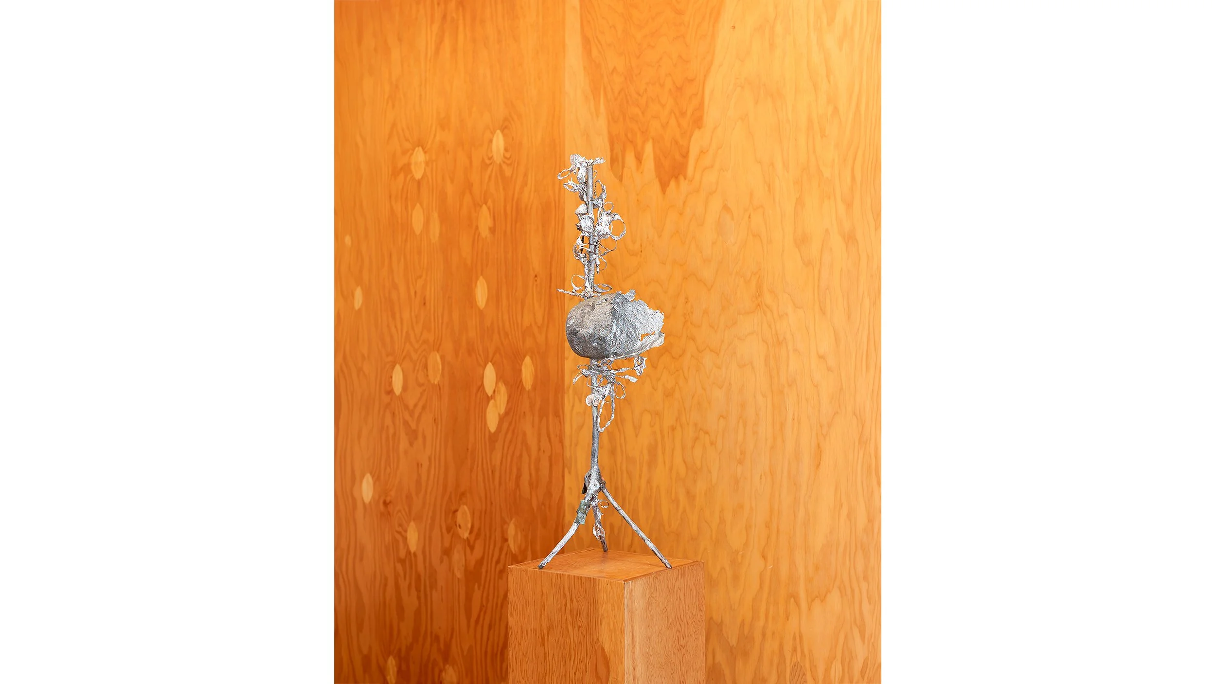 Yasue Maetake Three-Legged Idols gallery installation: single silver three-legged sculpture with rounded central form standing on wooden pedestal against plywood wall