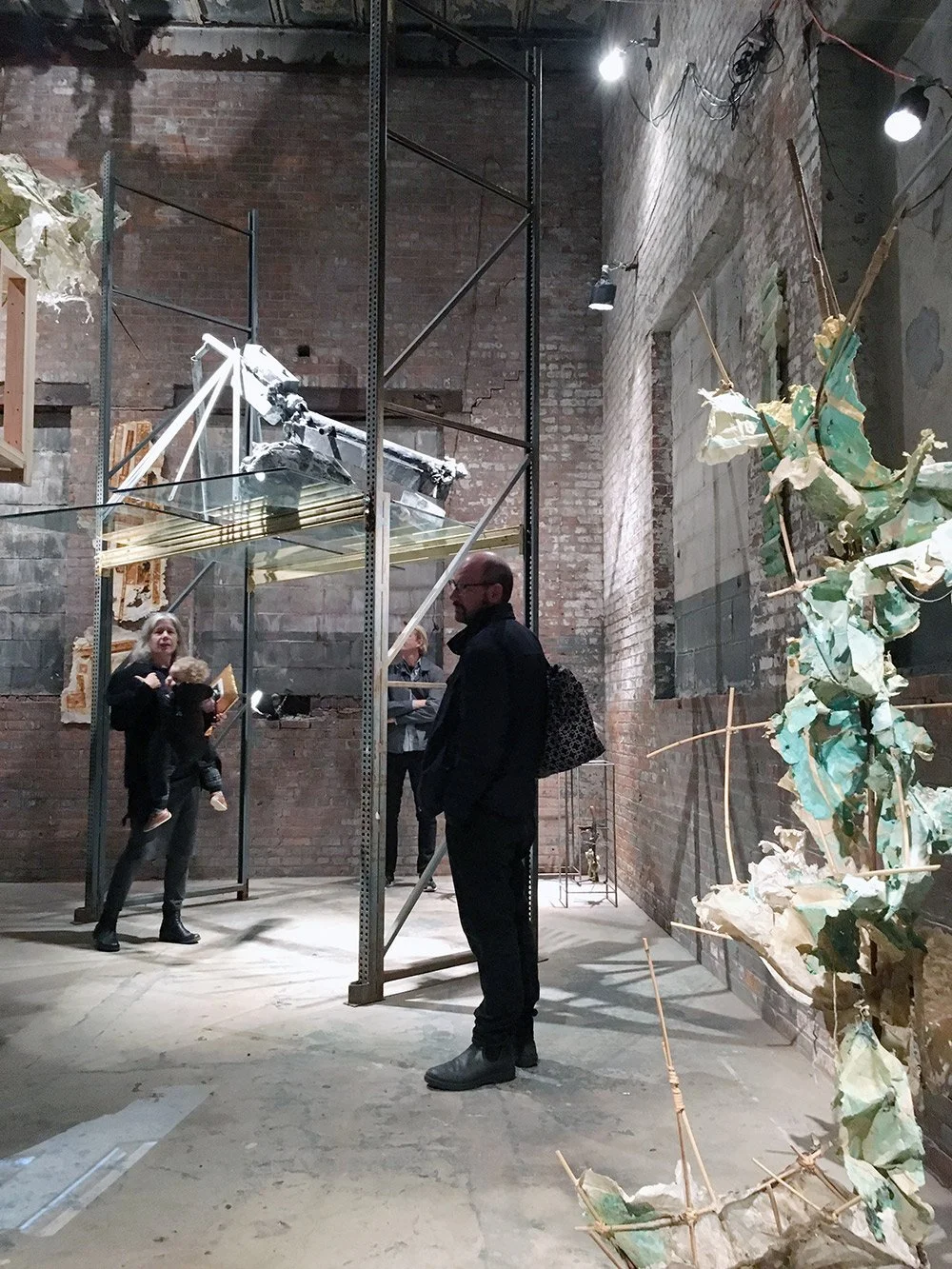 People viewing art installations in an industrial-style gallery with brick walls and exposed pipes. One woman holds a child, while others observe various sculptures and pieces on display.