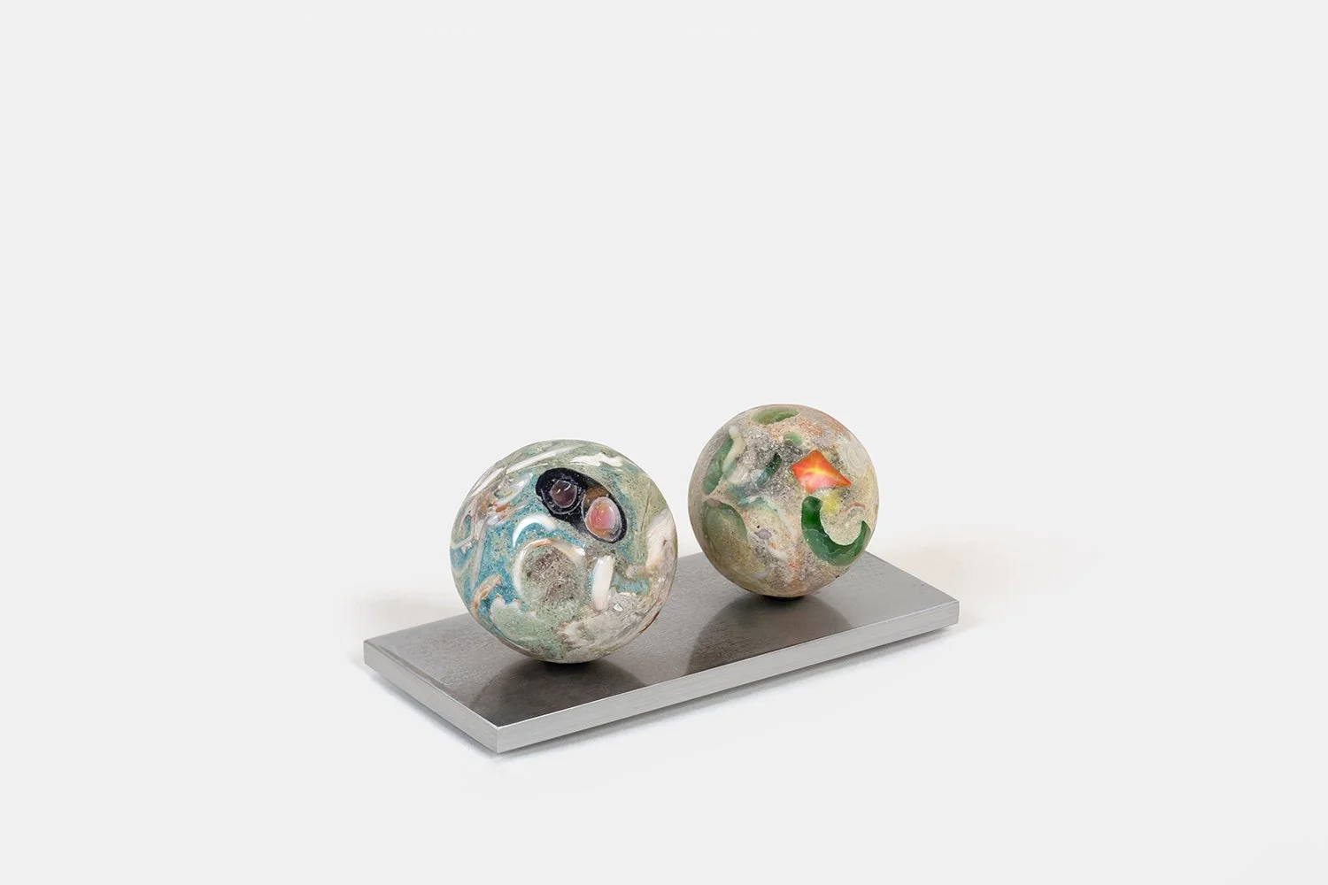 Yasue Maetake Orb Materia—Binoculars, 2026. Two spherical forms on a shared rectangular aluminum base, the left with circular black and white elements, the right with a bright orange fragment and green accents.