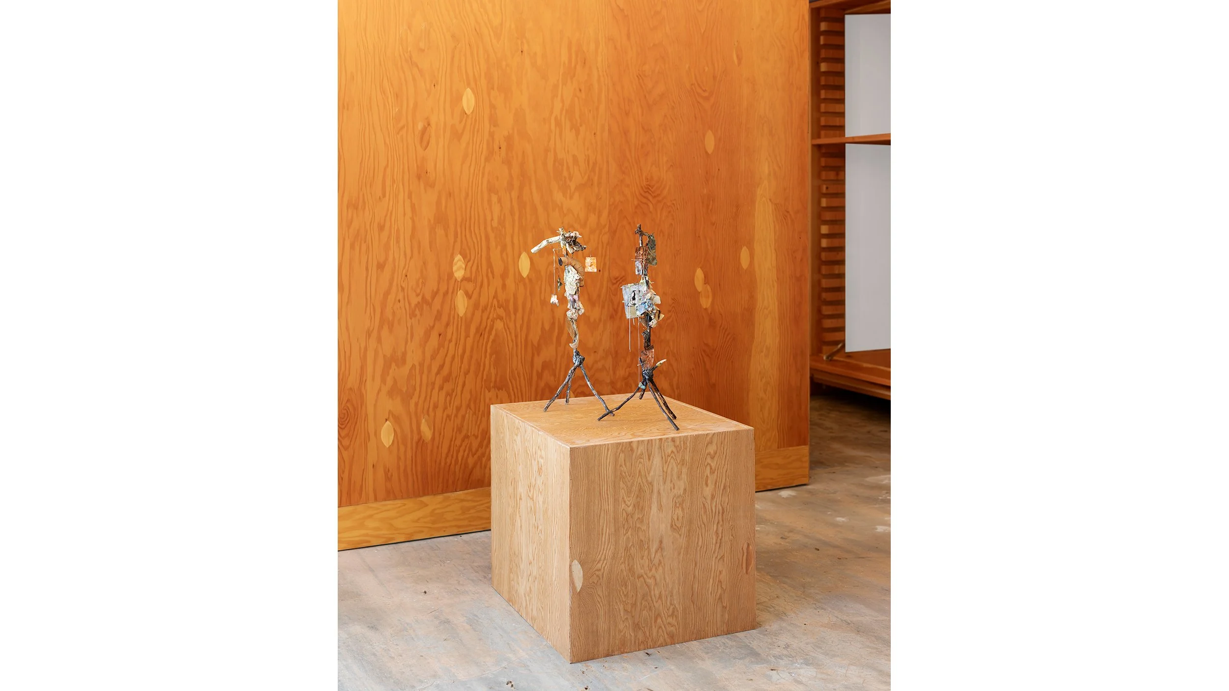 Yasue Maetake Three-Legged Idols gallery installation: two small three-legged sculptures standing side by side on a single wooden cube pedestal against plywood wall