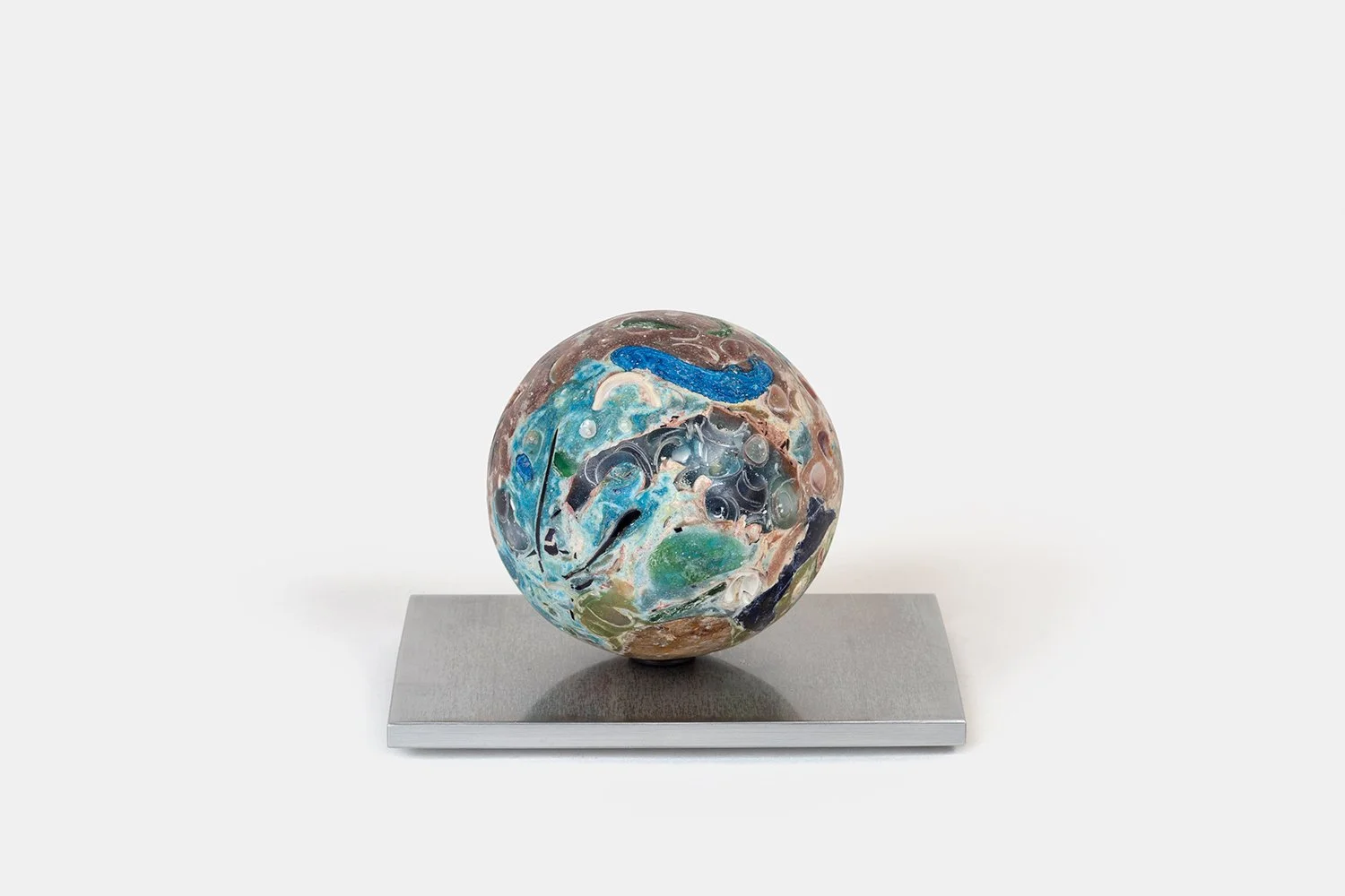 Yasue Maetake Orb Materia - Photosynthesis, 2026. Spherical form with dense blue, green, and brown mineral surfaces containing shell fragments and organic textures, resting on a square aluminum base.