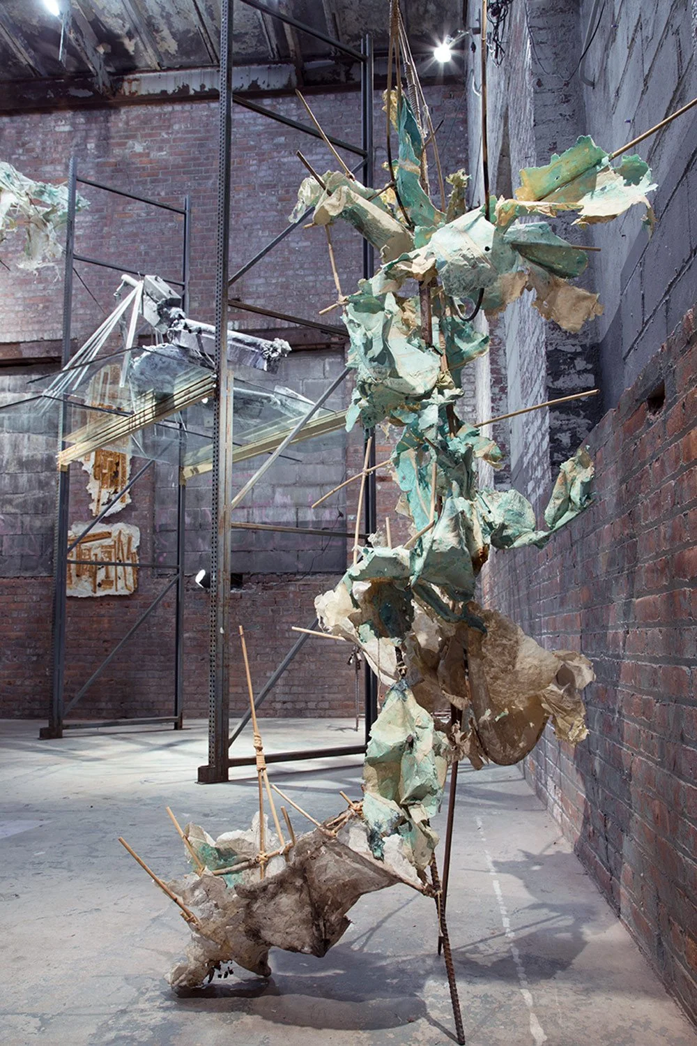 Mixed media art installation featuring a sculpture made of crumpled and torn paper, sticks, and a tree branch, displayed in an industrial space with brick walls and metal shelving.