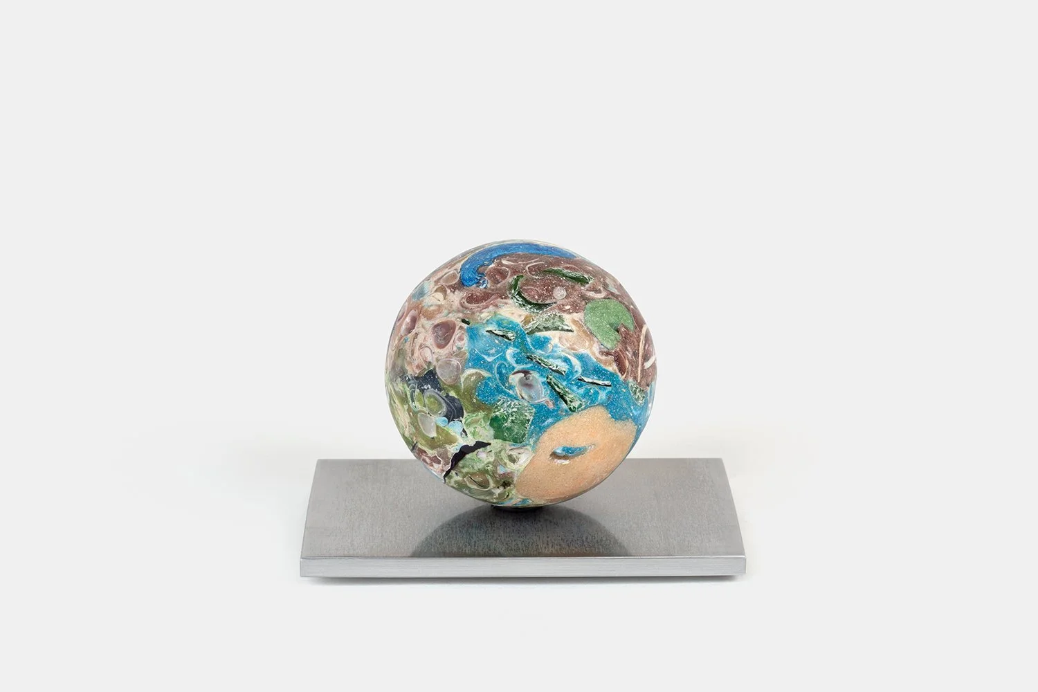 Yasue Maetake Orb Materia - Photosynthesis, 2026. Spherical form with dense green, blue, and brown mineral surfaces containing shell fragments, and a soft light beige area on the lower left that has an irregular, landmass-like shape, resting on a squ