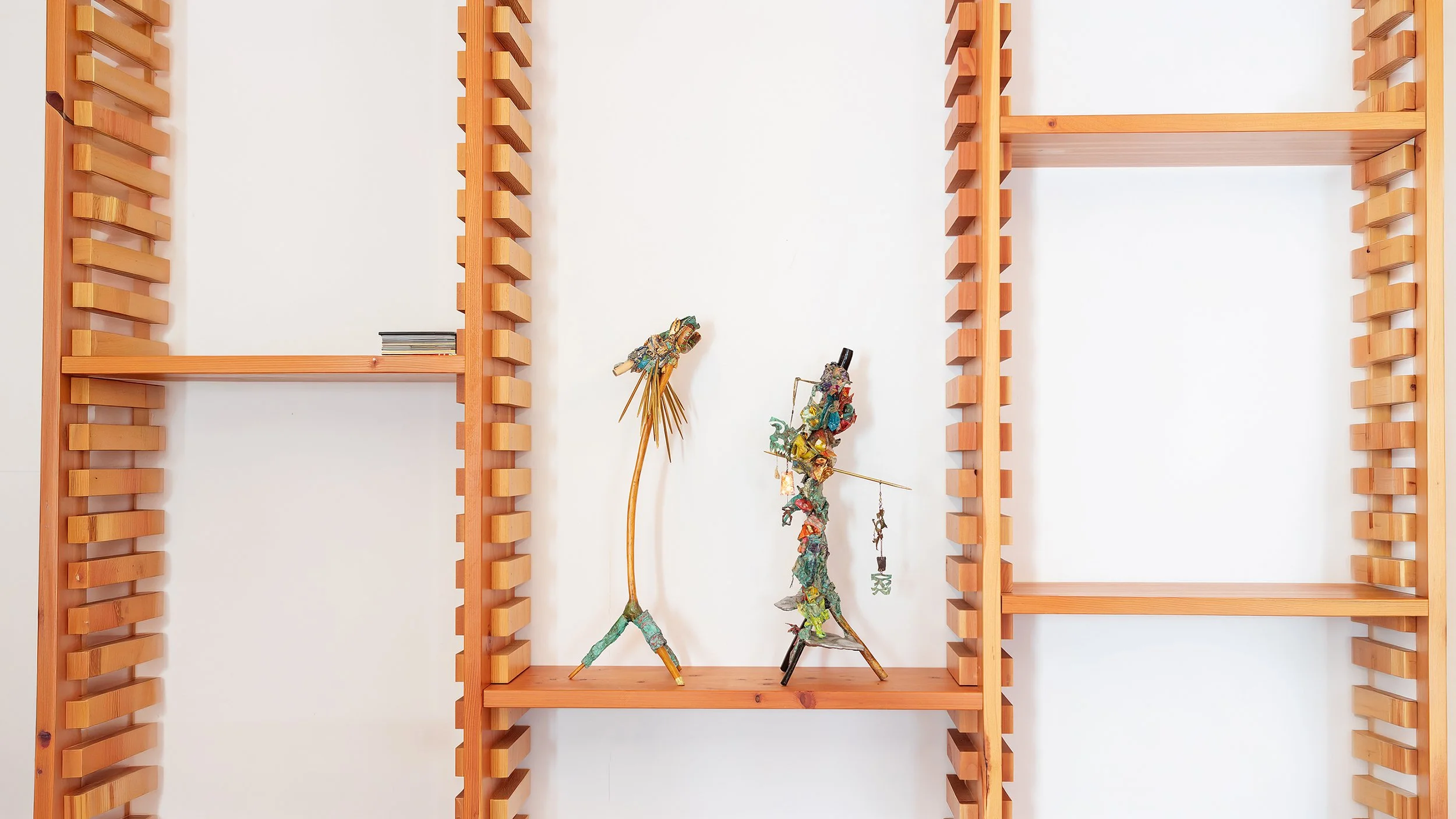 Yasue Maetake Three-Legged Idols detail: two small three-legged sculptures on wooden shelf, one tall thin yellowish form and one denser multicolored form with hanging elements