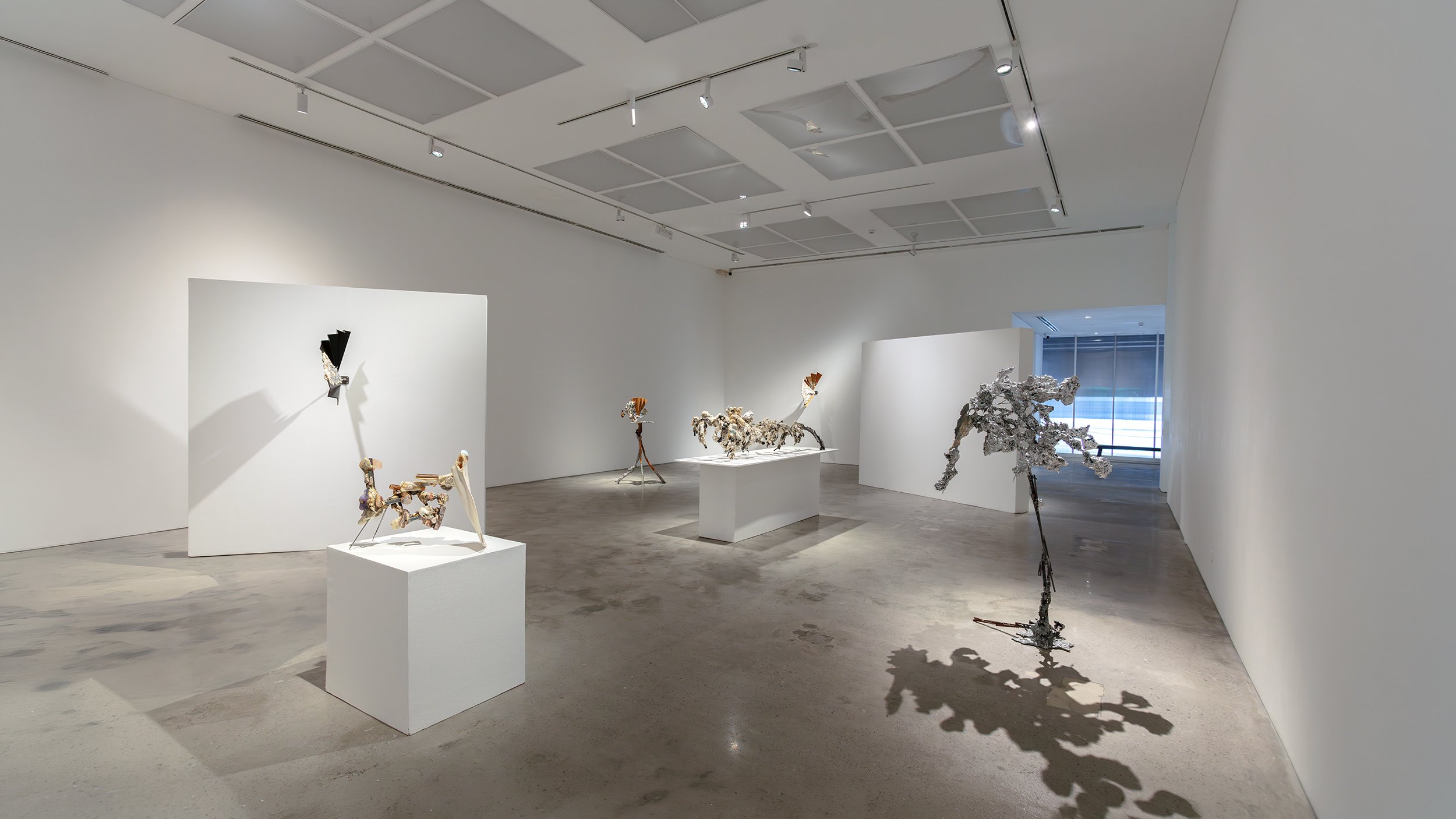 Yasue Maetake Mare Fecunditatis wide gallery view: black fan-shaped wall-mounted left, small orange tripod piece center left, long pedestal with multicolored dangling forms center, tall silvery tree-like on floor right, blinds window background