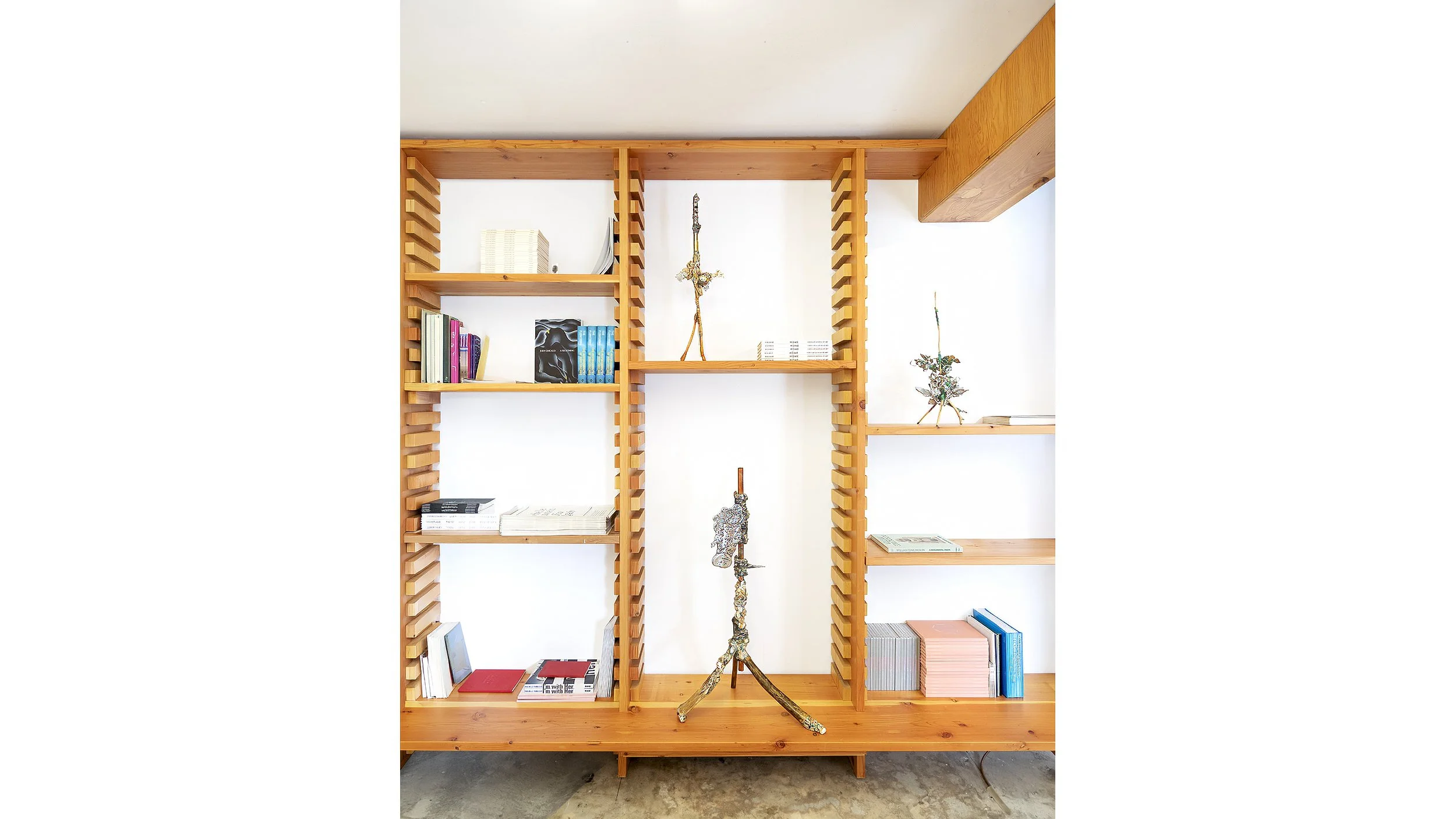 Yasue Maetake Three-Legged Idols gallery installation: wooden bookshelf section with several small three-legged sculptures placed among books on multiple shelves