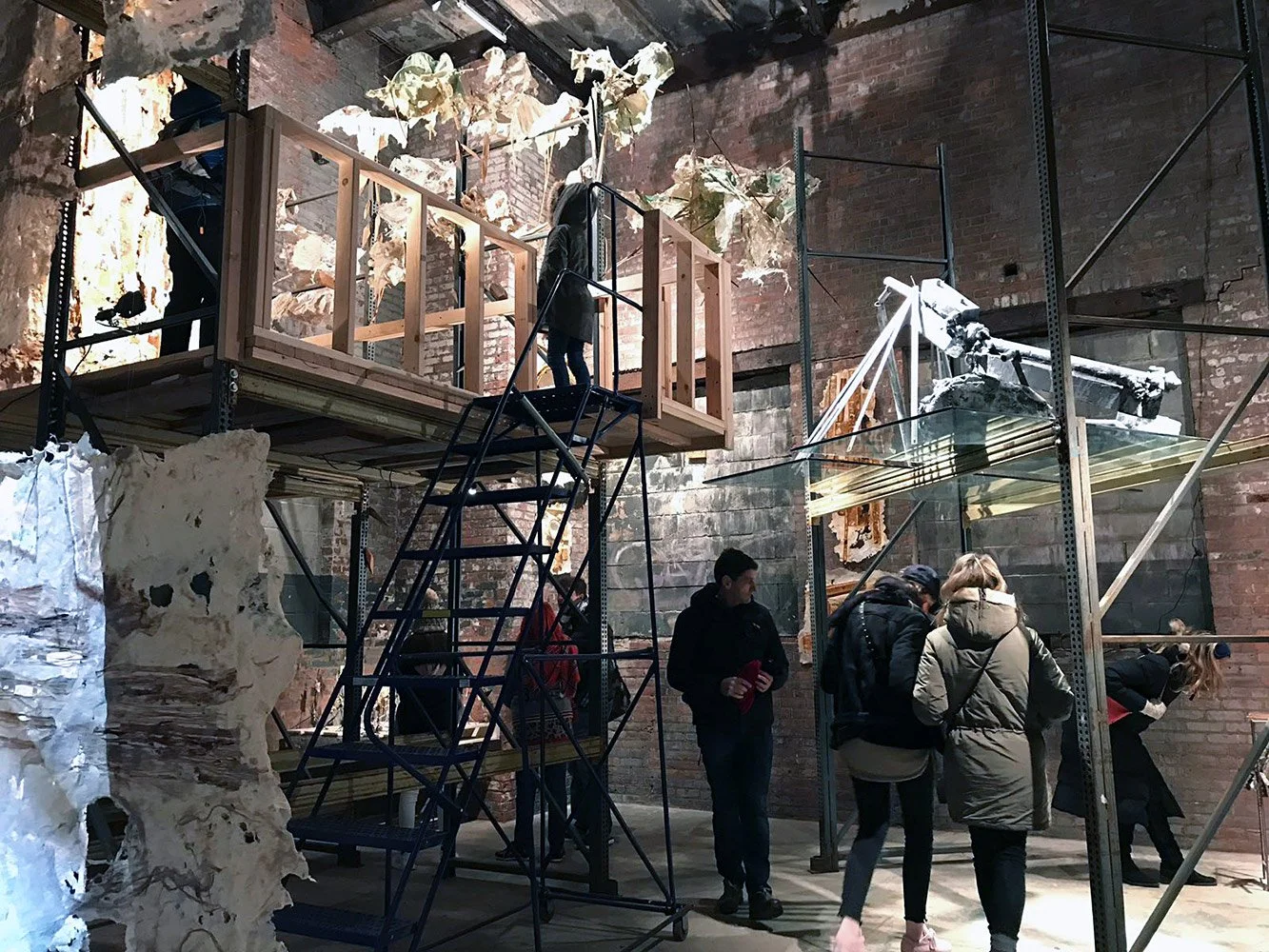 People observing an art installation in an industrial-style gallery with exposed brick walls, scaffolding, and mixed-media sculptures.
