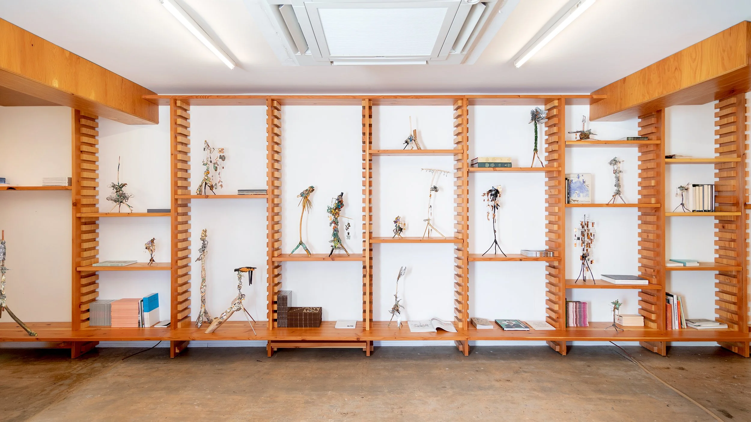Yasue Maetake Three-Legged Idols gallery installation: wide view of exhibition library wall filled with small three-legged sculptures placed among books and objects in white gallery space