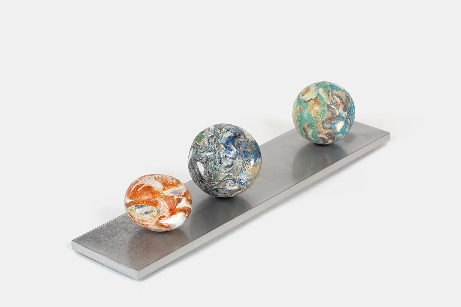 Yasue Maetake Orb Materia—Tangerine PCB, 2026. Three spherical sculptures on a long rectangular aluminum base, viewed from a higher angle showing orange, blue-gray, and turquoise mineral surfaces with embedded fragments.