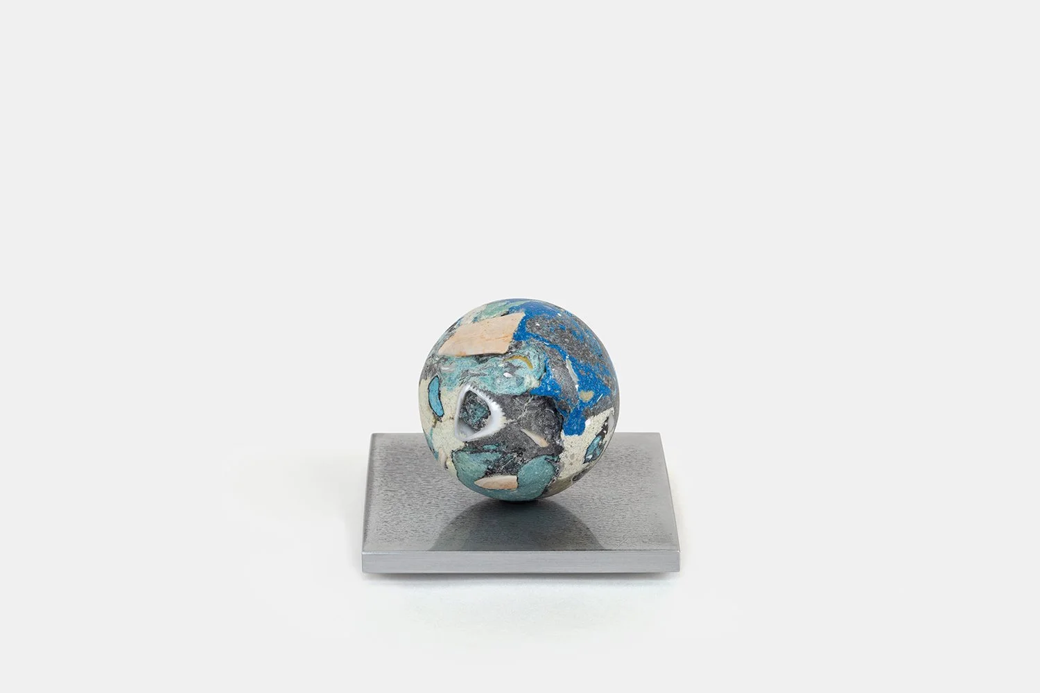 Yasue Maetake Orb Materia—Blue Float, 2026. Compact spherical sculpture with deep blue and dark mineral surfaces containing beige-white shell fragments, resting on a square aluminum base.