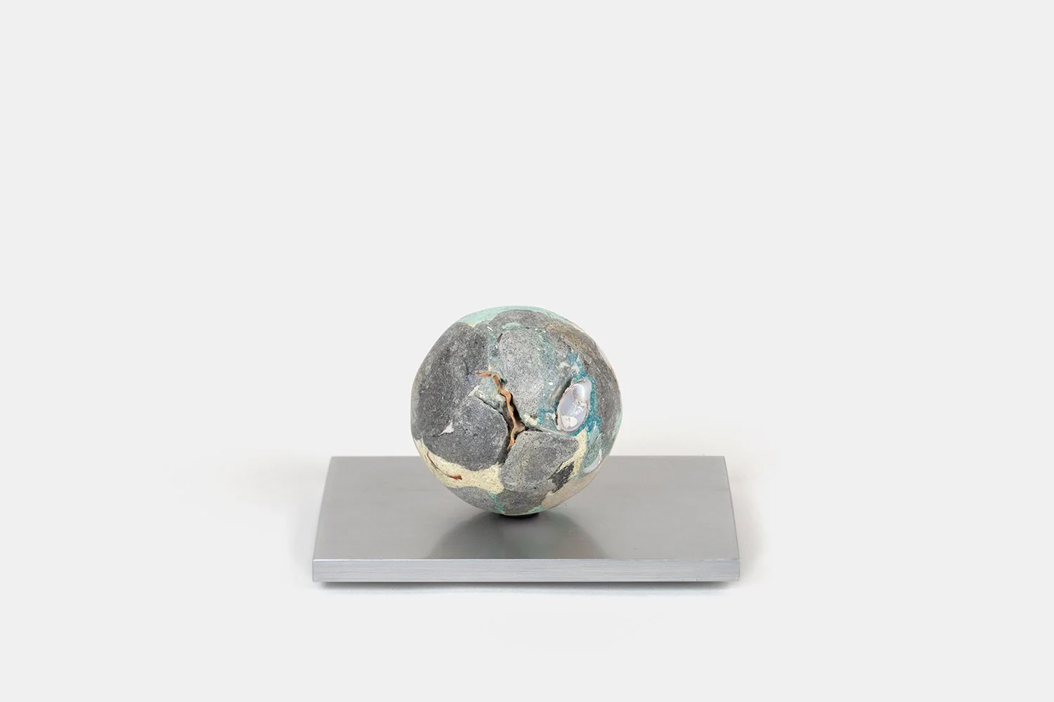 Yasue Maetake Orb Materia—Air Current, 2026. Spherical form with dark gray cracked mineral surface, soft turquoise accents, and a small white shell fragment, resting on a square aluminum base.