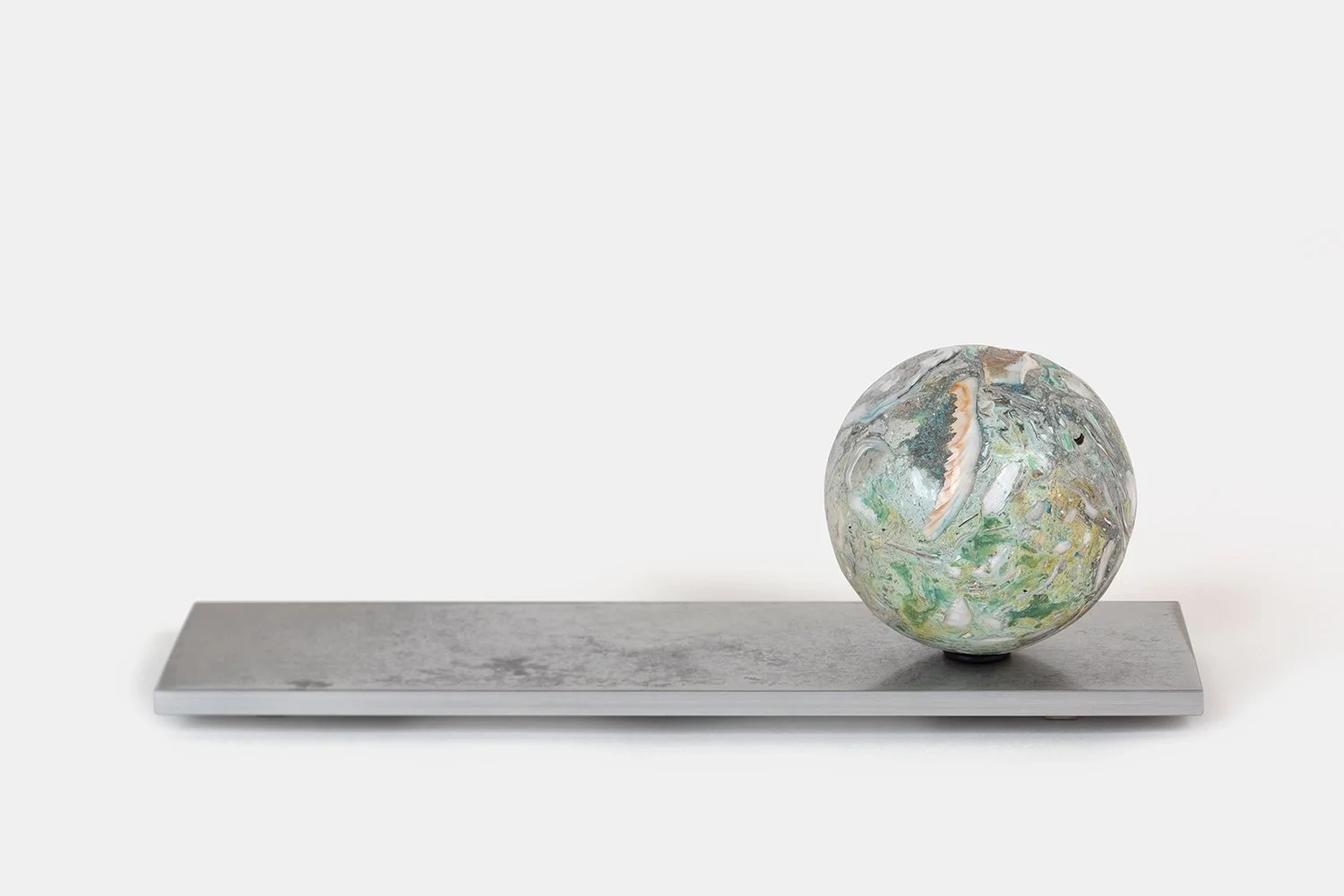 Yasue Maetake Orb Materia—Chlorophyll, 2026. Spherical form with soft green, white, and beige mineral surfaces containing shell fragments, positioned on the right side of a long rectangular aluminum base.